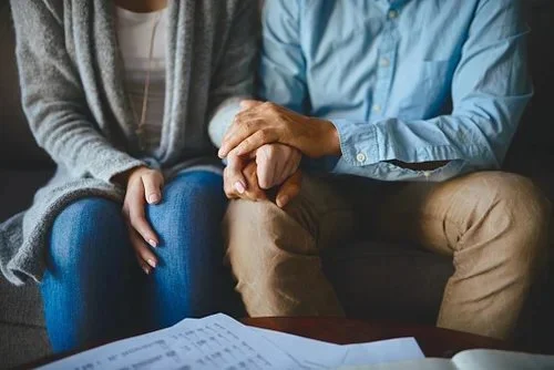 A person in a gray sweater holding hands with a person in a blue shirt and tan pants, sitting close together, with papers on a table in front of them.