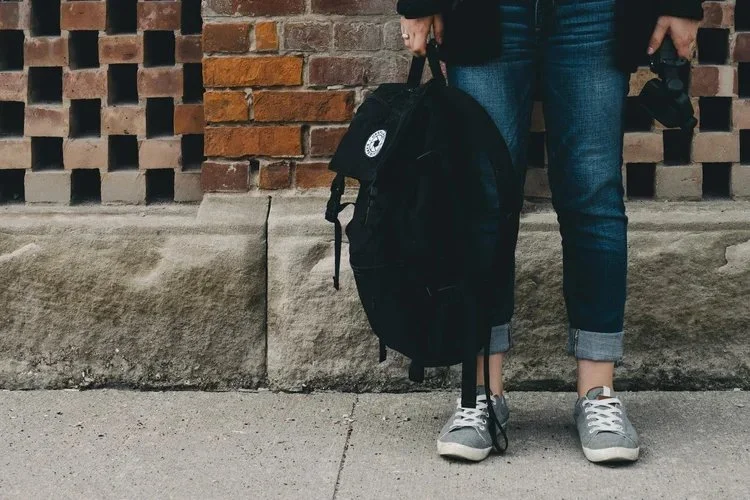 Person standing on a sidewalk holding a black backpack, wearing sneakers, rolled-up jeans, and a dark jacket, with a brick wall in the background.
