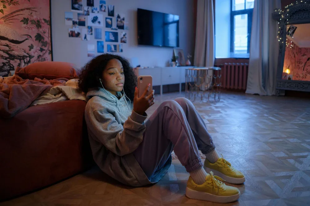 A young woman with curly hair sitting on the floor, leaning against a couch, looking at her phone, in a cozy, well-decorated living room.