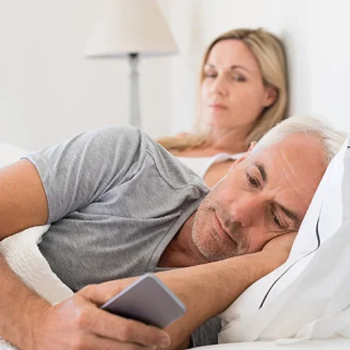 A middle-aged man is lying in bed, looking at his smartphone, while a woman is sitting in the background, appearing to be listening or watching.