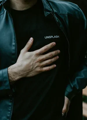 Person wearing a black shirt with the word 'UNSPLASH' on it, partially covered by a dark jacket, with hand placed over chest.