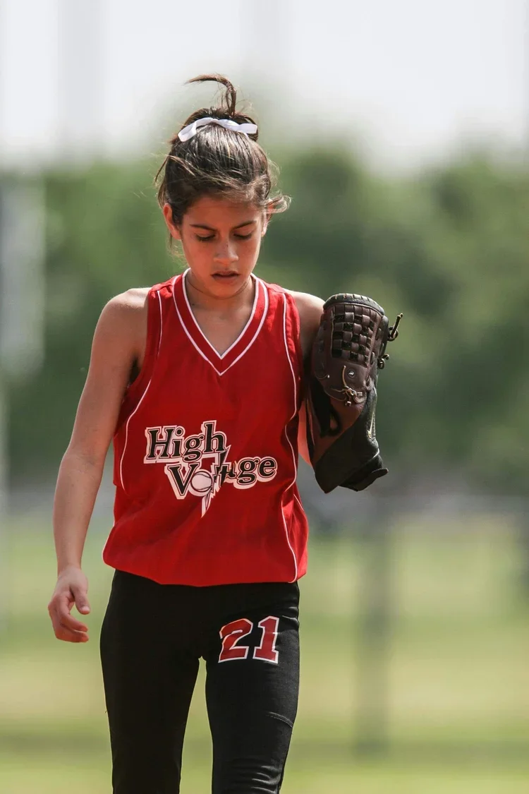 Young female softball player in red jersey with 'High Voltage' logo, black pants with number 21, holding a glove, looking down, outdoors.