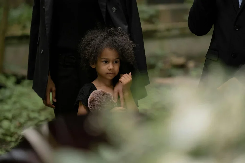 Young girl with curly hair holding a teddy bear, standing among adults in formal attire, outdoors.