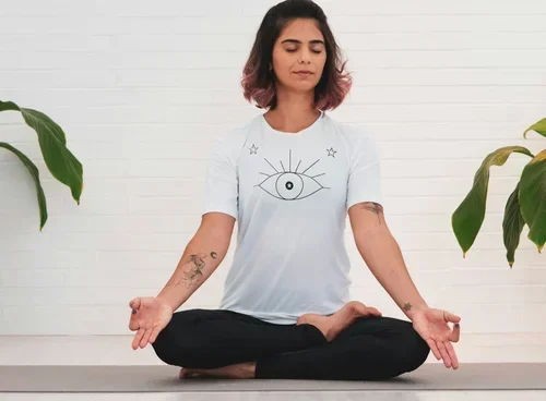 Young woman practicing meditation in a seated cross-legged pose with hands resting on her knees, wearing a white T-shirt with an eye and star design, outdoors with plants and a white wall in the background.