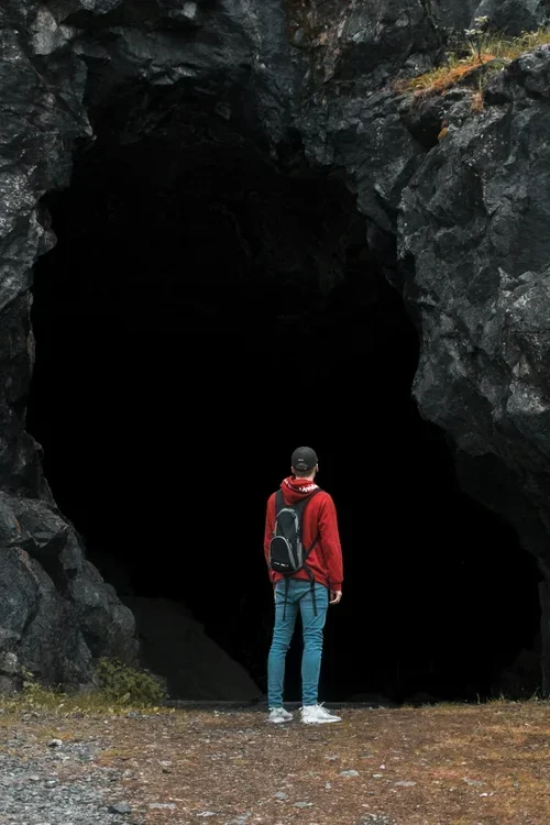 A person in a red jacket and blue jeans stands in front of a dark, large cave opening surrounded by rocky terrain.