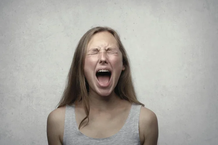 Young woman with long brown hair wearing a gray tank top, eyes shut tightly, mouth open as if yawning or shouting, standing against a plain beige wall.