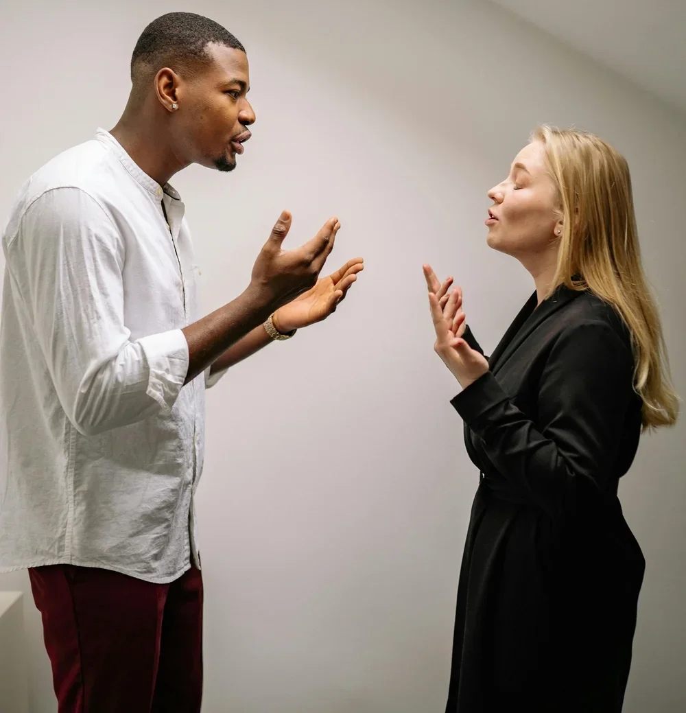 Two people, a man and a woman, engaging in a serious conversation indoors against a plain wall. The man appears to be explaining something, using hand gestures, and the woman is listening attentively with her eyes closed, also using hand gestures.