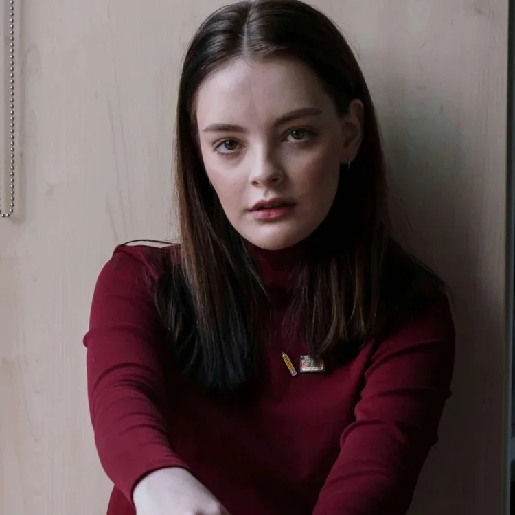 A young woman with long dark brown hair wearing a red top standing against a plain wood wall.
