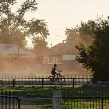A person riding a bicycle on a paved path with a fence in a suburban neighborhood during sunset.