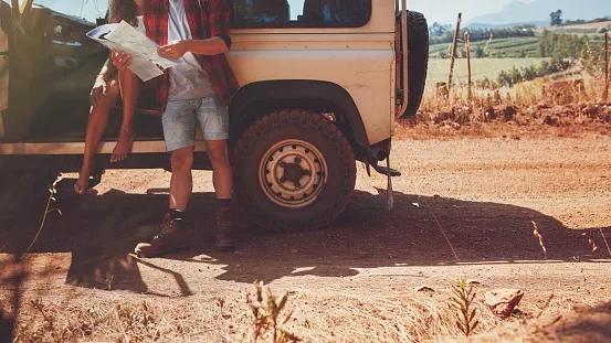 Two children standing beside a beige off-road vehicle on a dirt road, holding papers or maps. The landscape is rural, with fields and trees in the background.
