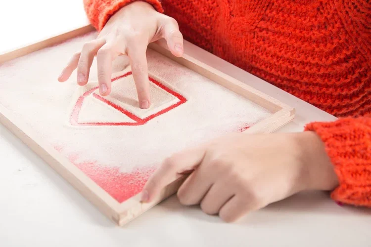 Child using their finger to draw a red checkmark on a white sand art board with a wooden frame, wearing an orange sweater.