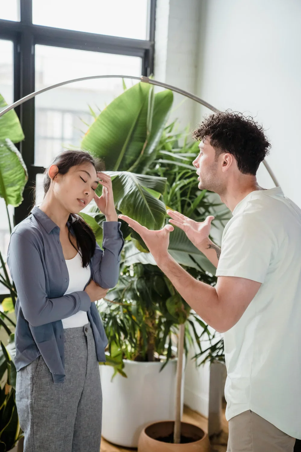 A woman appears distressed, holding her head, as a man gestures with his hands in a confrontational manner in front of her, with large green plants and large windows in the background.