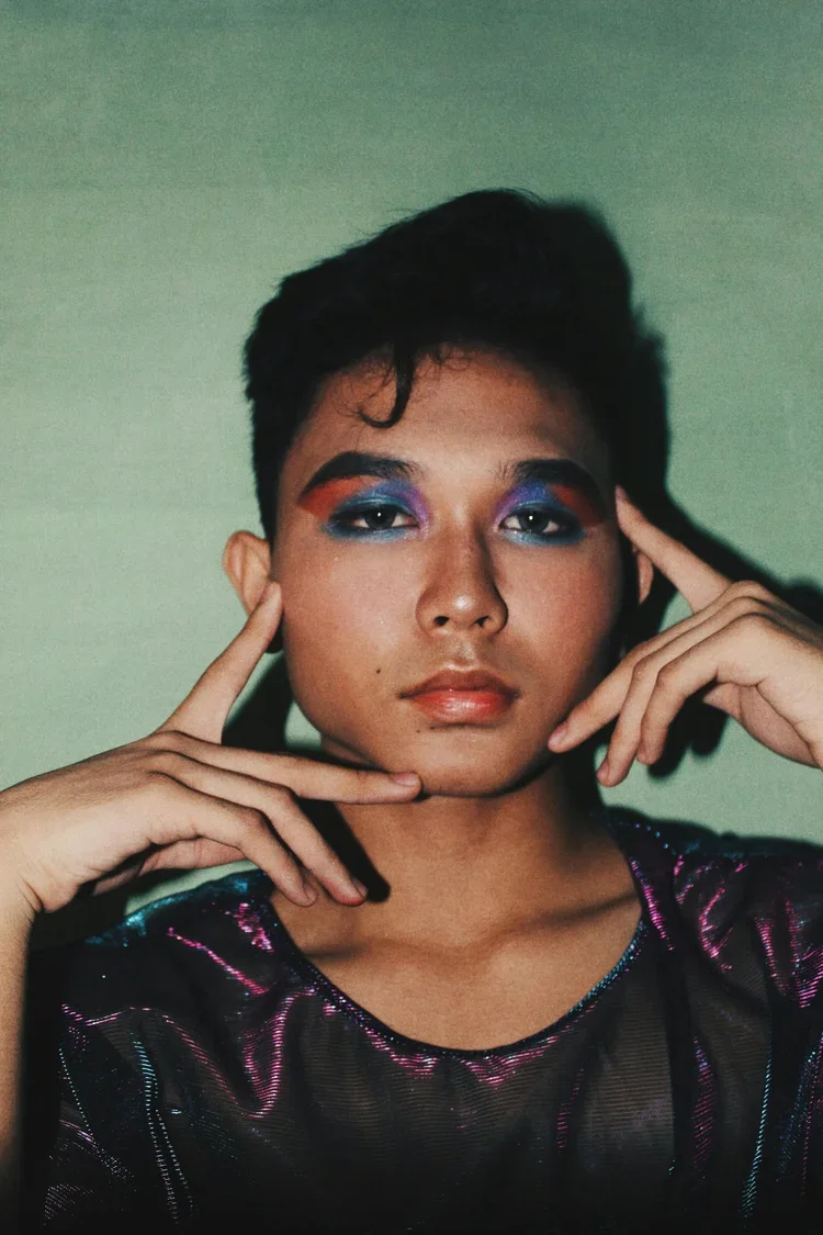 Portrait of a person with short dark hair and vibrant colorful makeup, wearing a shiny black top, posing with fingers framing their face.