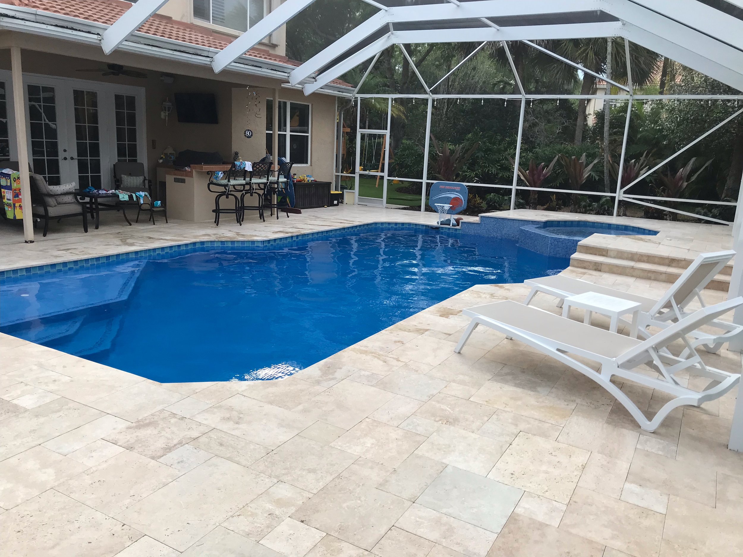 A backyard with a swimming pool, patio area with outdoor furniture, and two white lounge chairs on a beige stone deck. The area is enclosed in a screened structure with surrounding greenery.