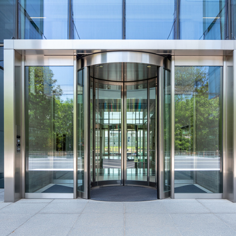 Modern glass building entrance with revolving door and surrounding glass walls, reflecting trees and the sky.