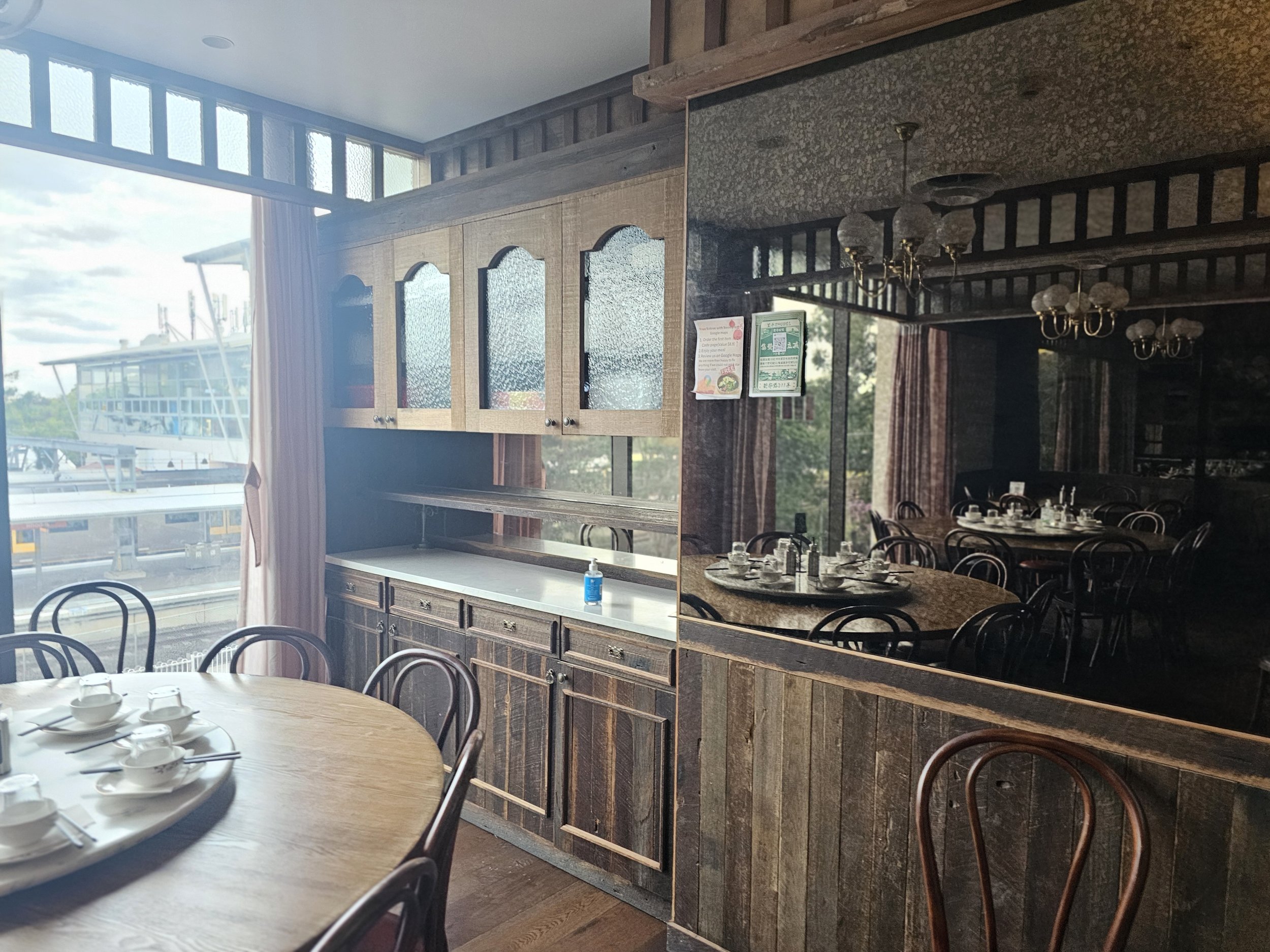 Interior of a restaurant with wooden cabinets, a dining table set with bowls and utensils, and a mirror reflecting a dimly lit dining area with multiple tables and chairs.