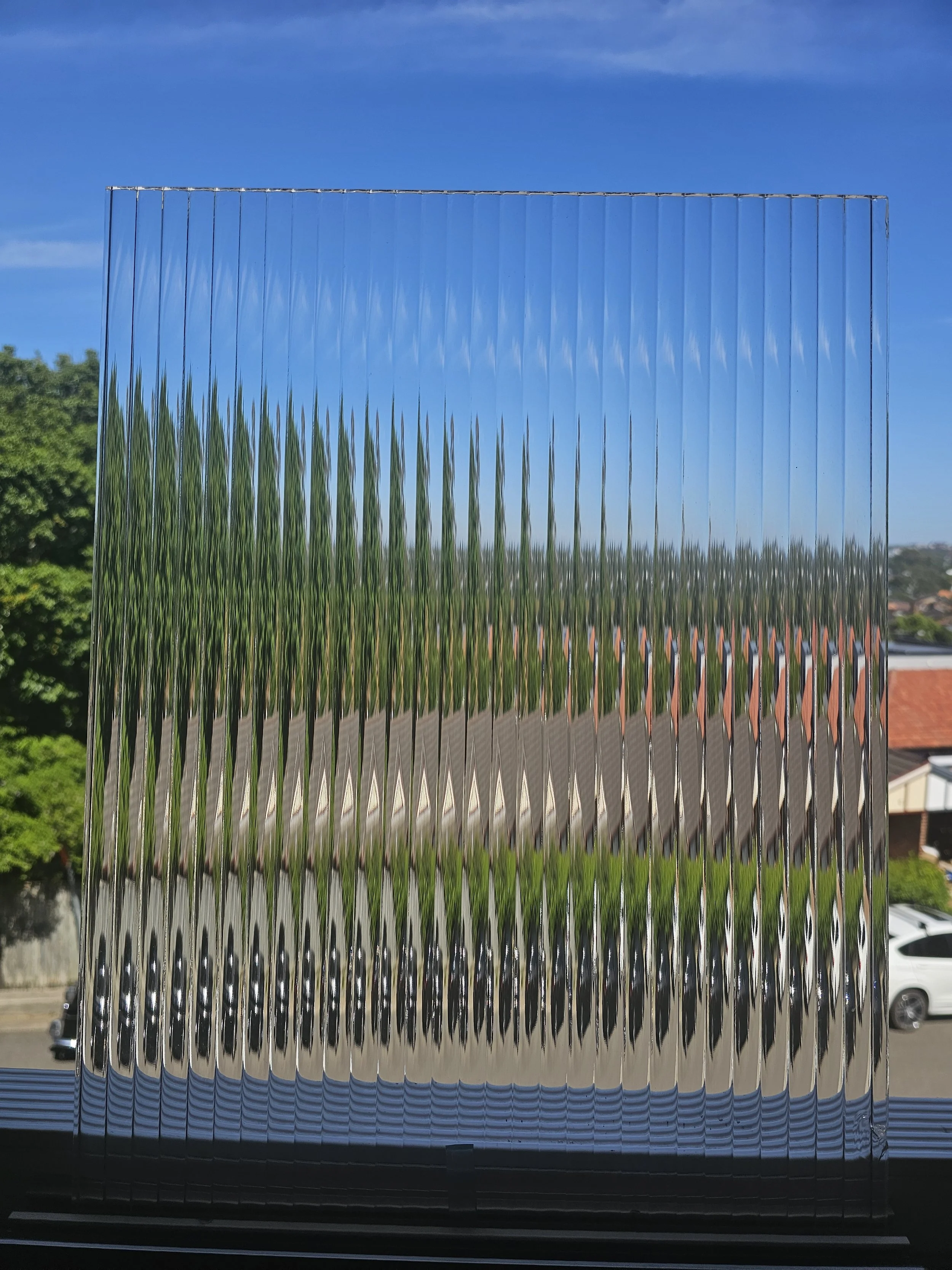 A glass panel with a diamond pattern placed on a window, reflecting a green trees, a building with a red roof, and a blue sky with some clouds outside.