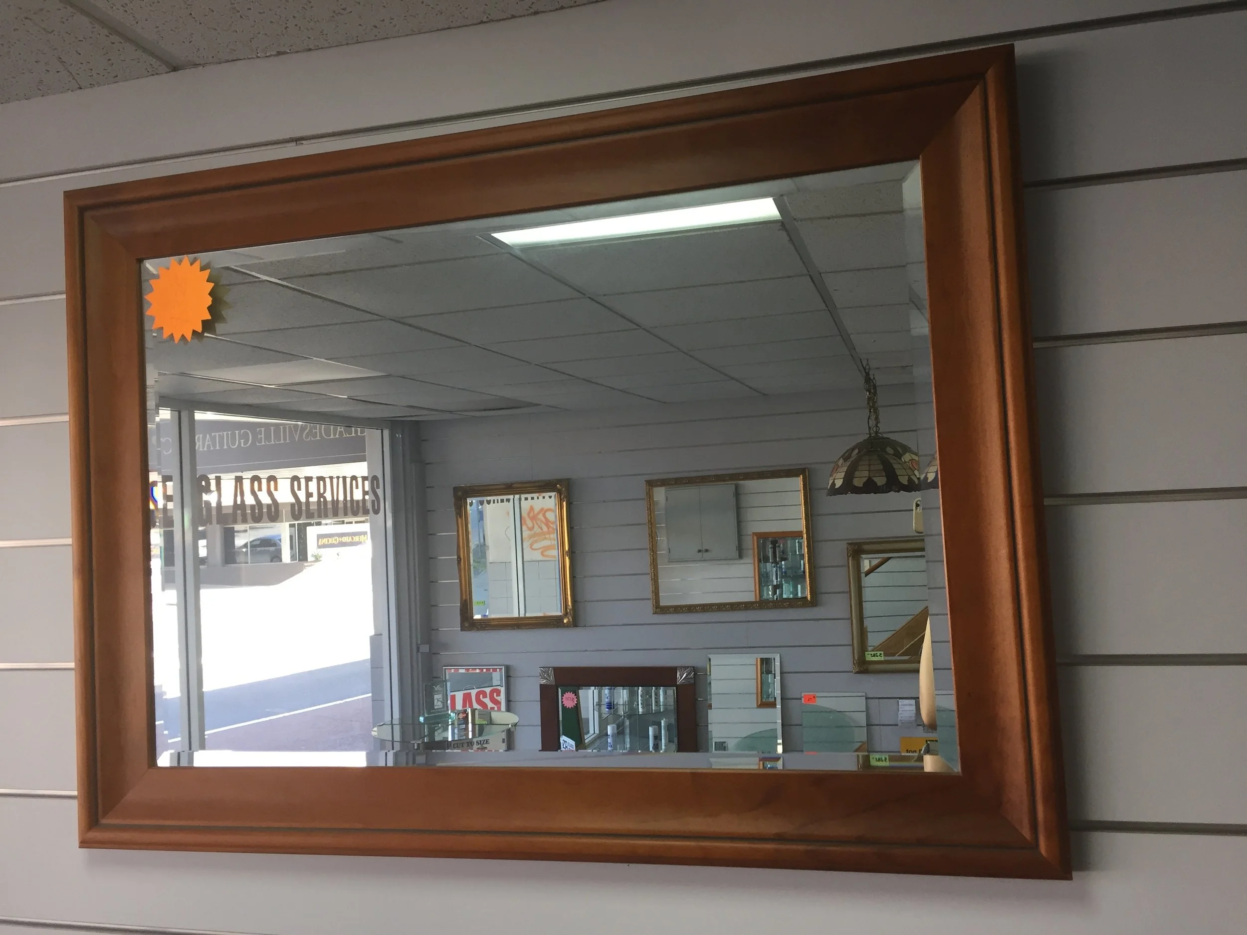 Interior of a glass shop showing various framed mirrors on a wall, a hanging lamp, and a display case, with a door and window showing outside signage reading 'Glass Services'.