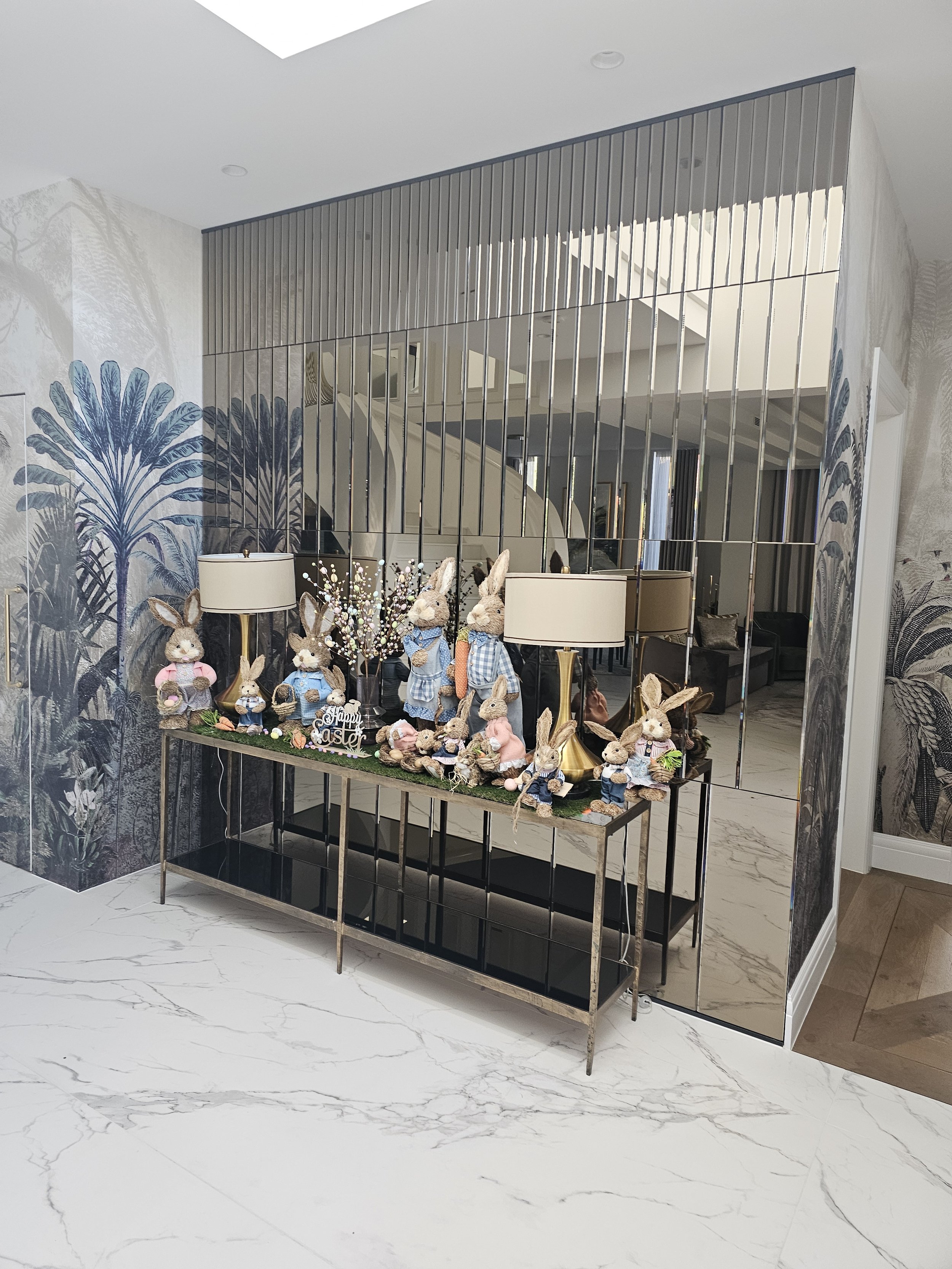 Decorative display with Easter-themed rabbit plush toys and figurines on a table, set against a mirror and floral patterned wallpaper wall.