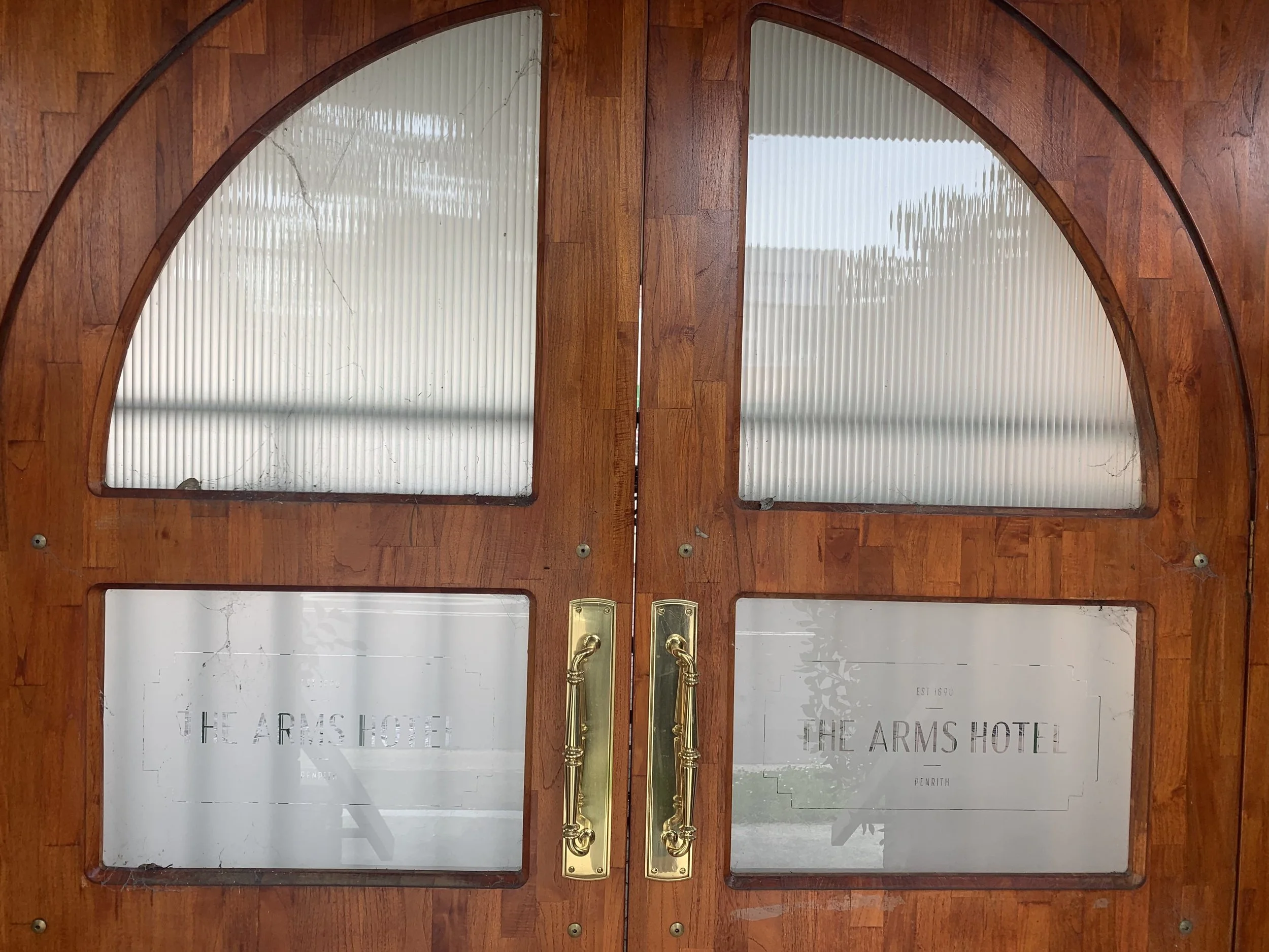 Wooden double door with glass panels, brass handles, and the text "The Arms Hotel Penrith" on the lower glass panels.