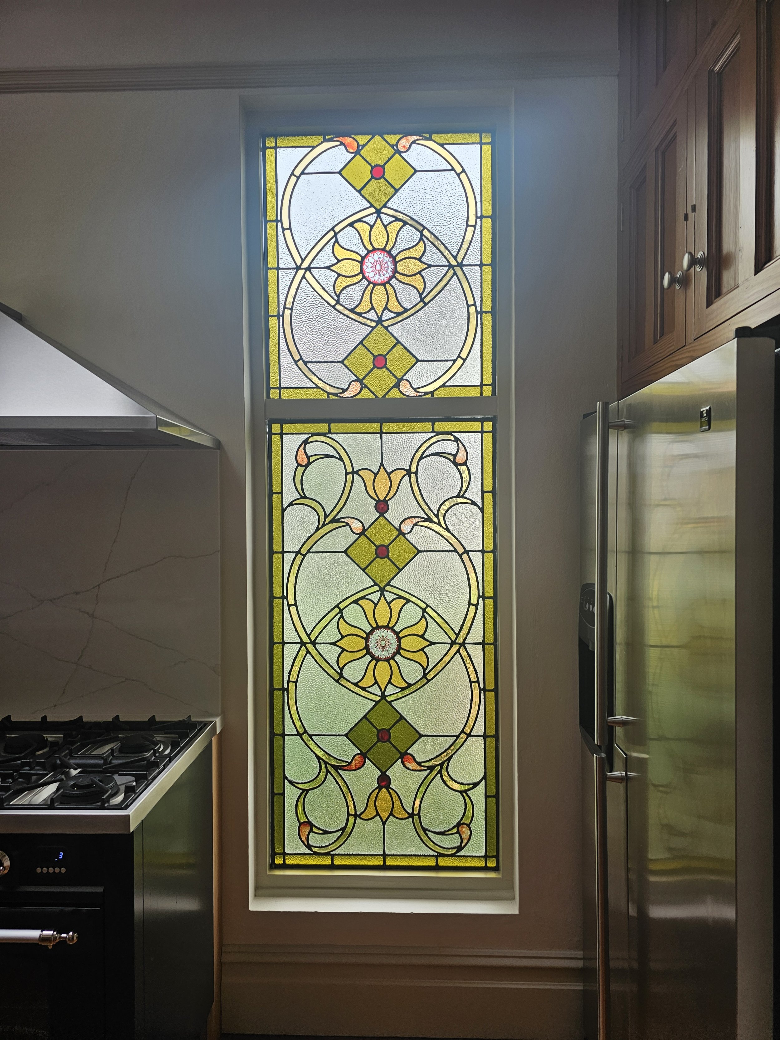Decorative stained glass window with geometric and floral patterns, situated between a kitchen stove on the left and a refrigerator on the right.
