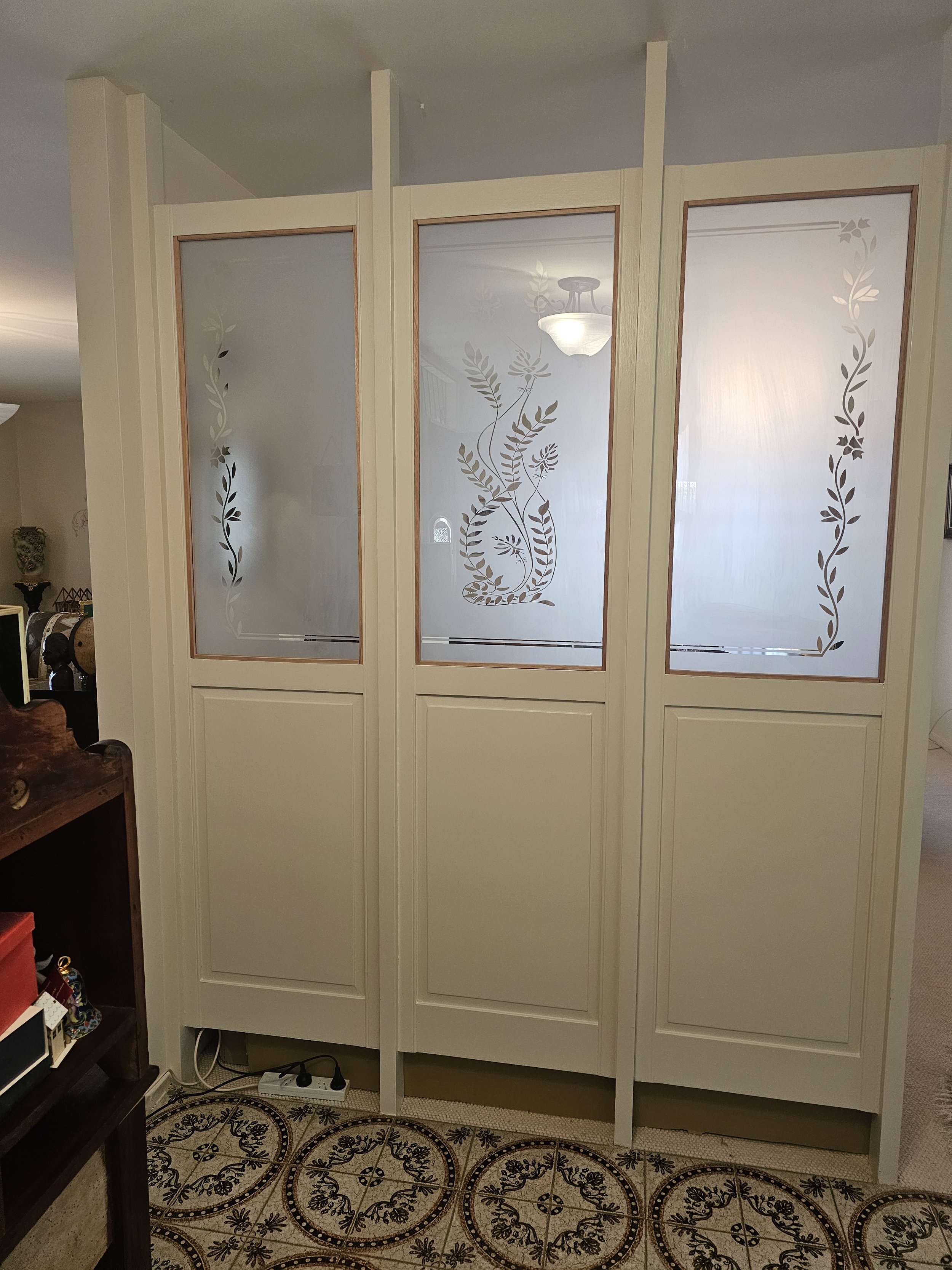 A decorative white room divider with three panels, each featuring frosted glass with floral and leaf patterns etched on it, standing on a patterned tile floor in a living room.