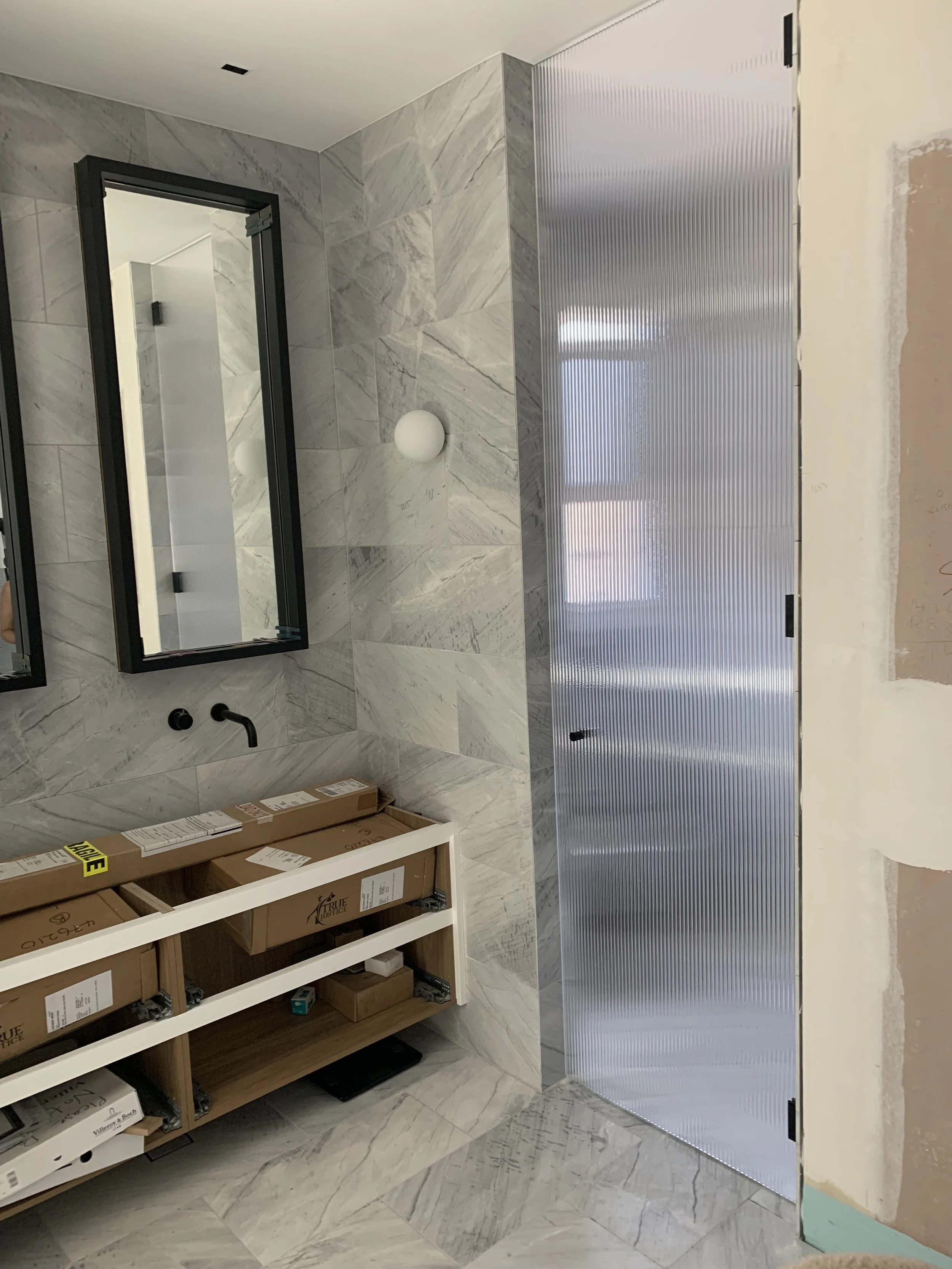 A bathroom under construction with marble tiles on the walls and floor, a black-framed mirror, a wall-mounted black faucet, and a partially assembled wooden vanity cabinet with boxes inside, next to a metal door with a textured surface.