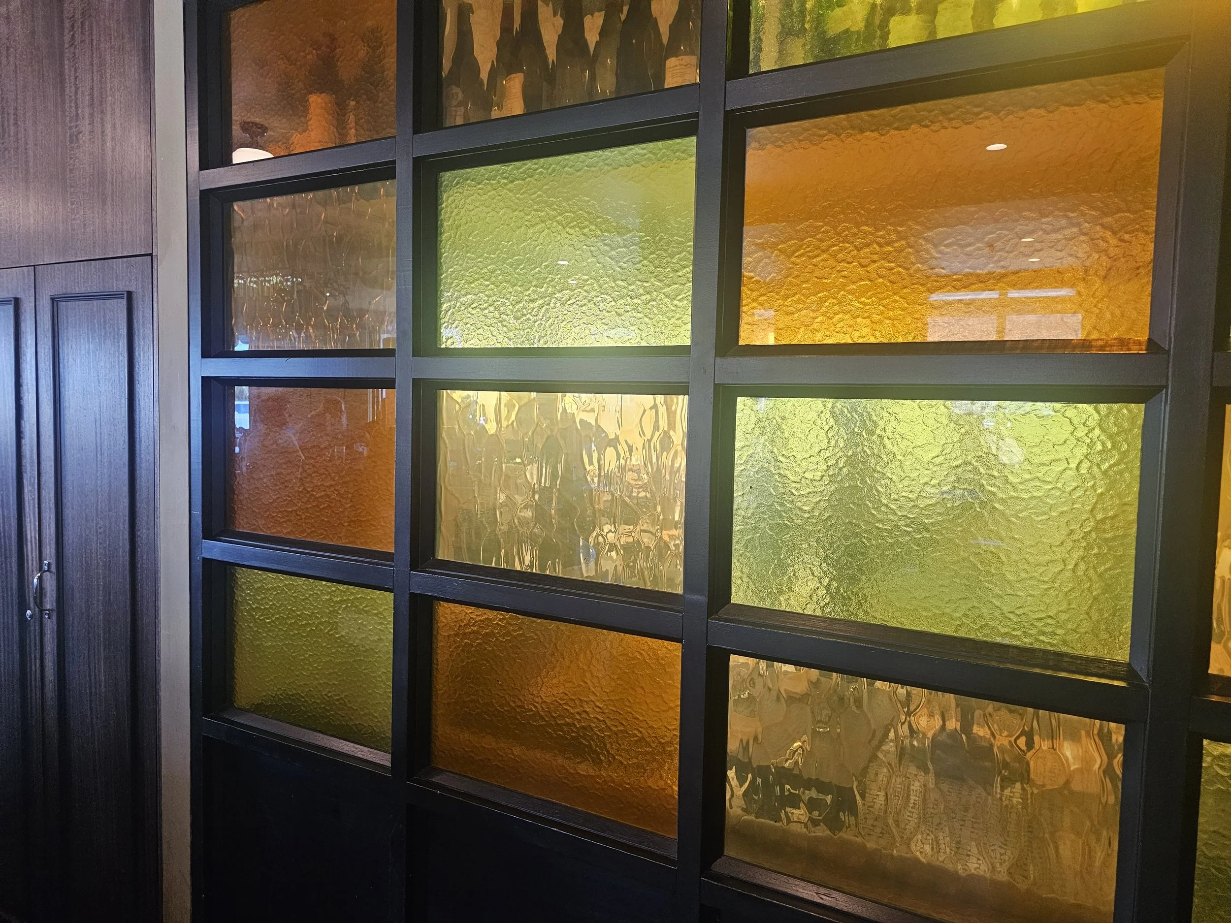 A black grid partition with textured glass panels in shades of yellow, green, and amber, mounted on a wooden wall.