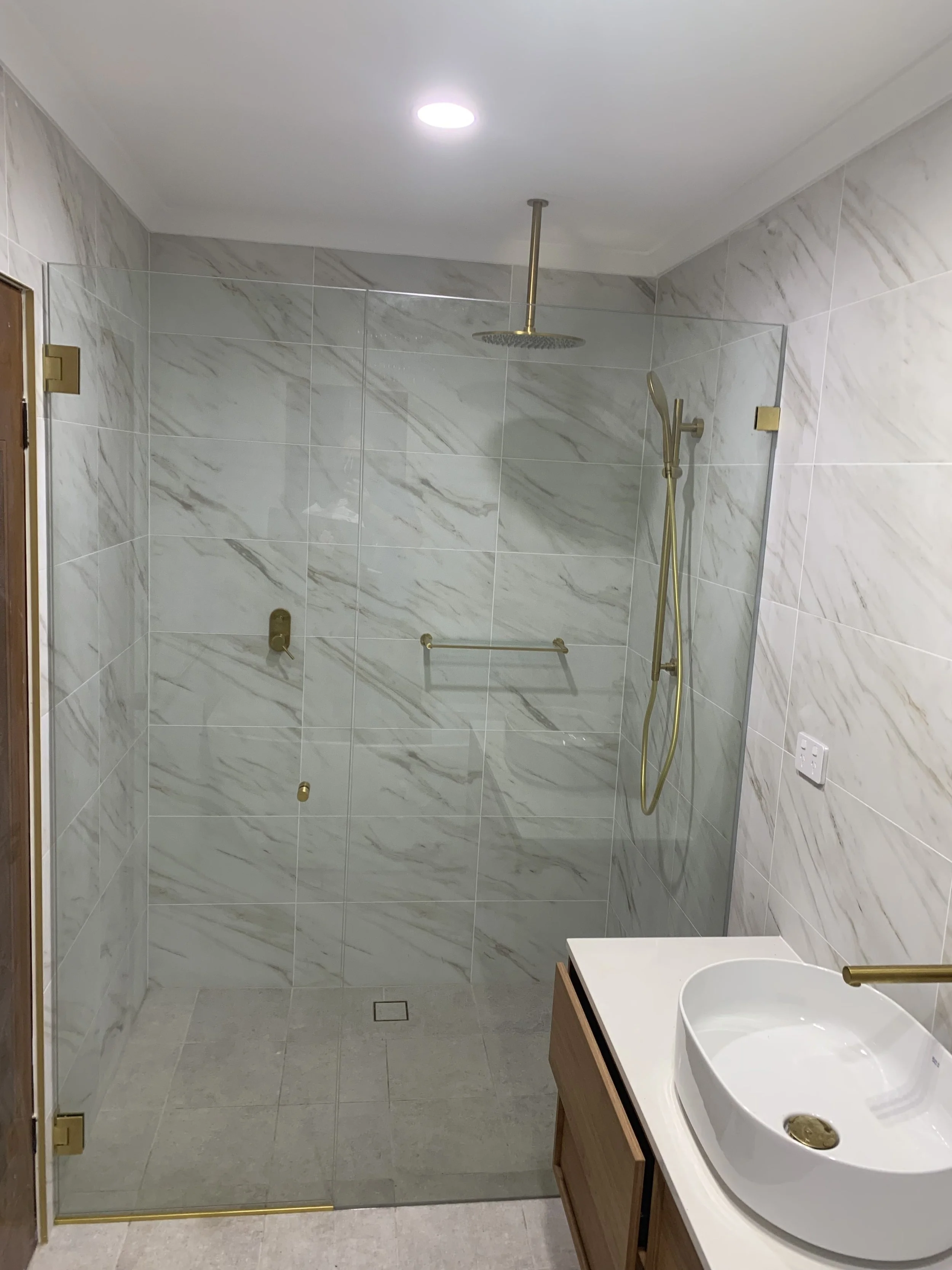 Modern bathroom with a walk-in shower enclosed by glass, featuring a rainfall showerhead and handheld shower fixture with gold accents. A wooden vanity with a vessel sink and gold faucet is visible to the right.