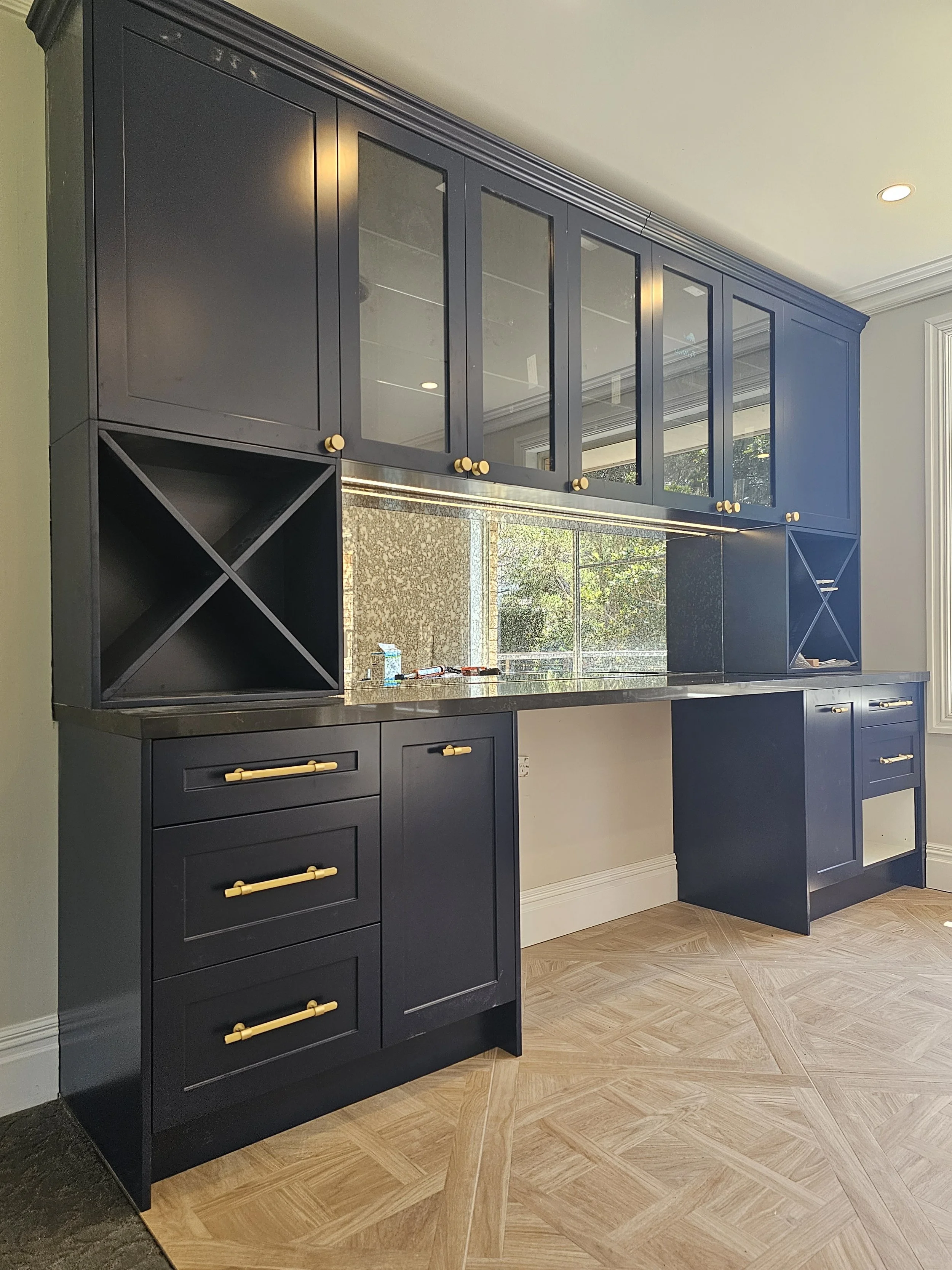 Open dark blue kitchen cabinet with gold hardware, glass-front upper cabinets, wine rack on both sides, and a reflection of a window in the backsplash mirror.