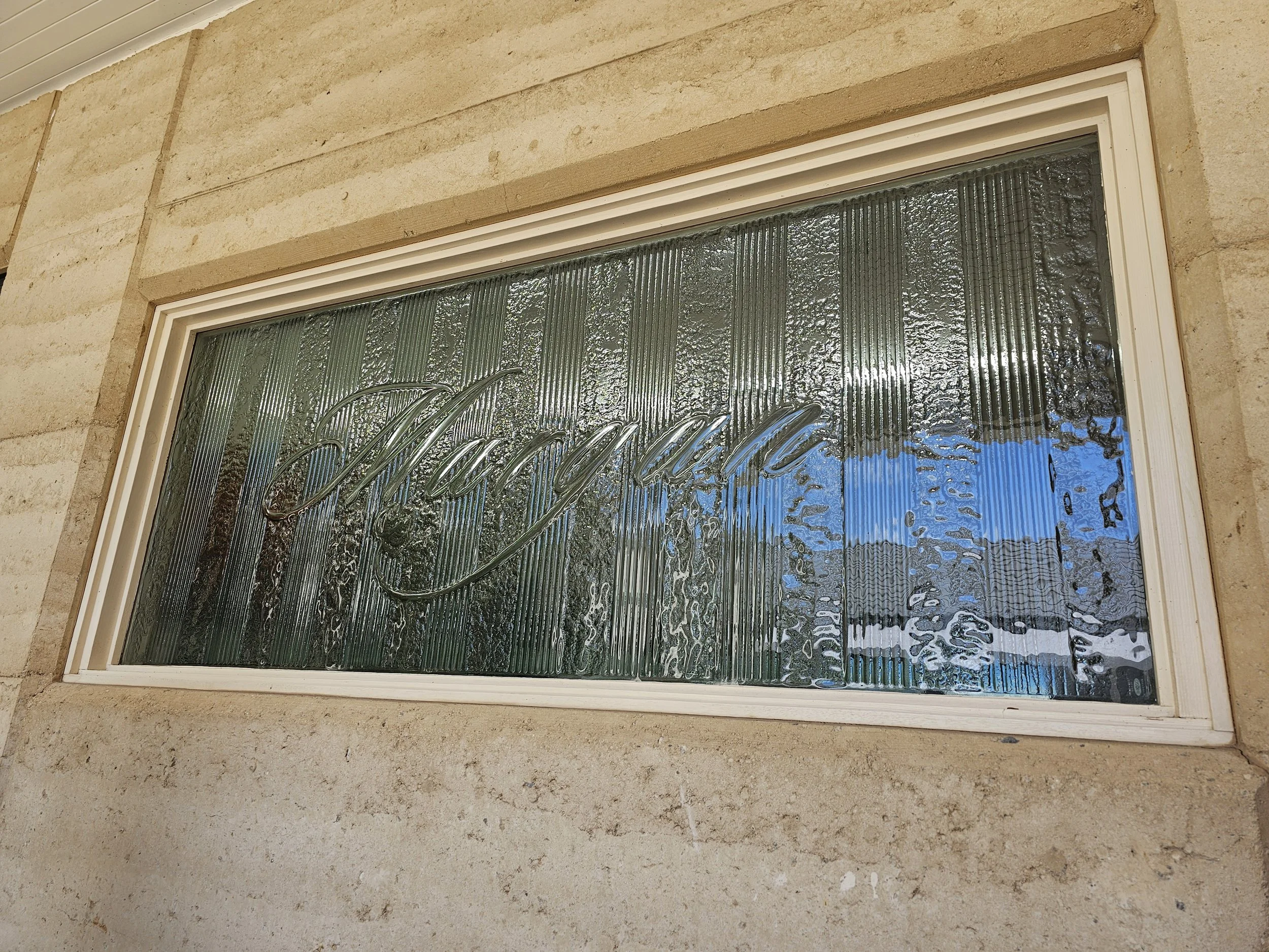 A rectangular glass panel with the word "Welcome" engraved in cursive, mounted on a beige brick wall.