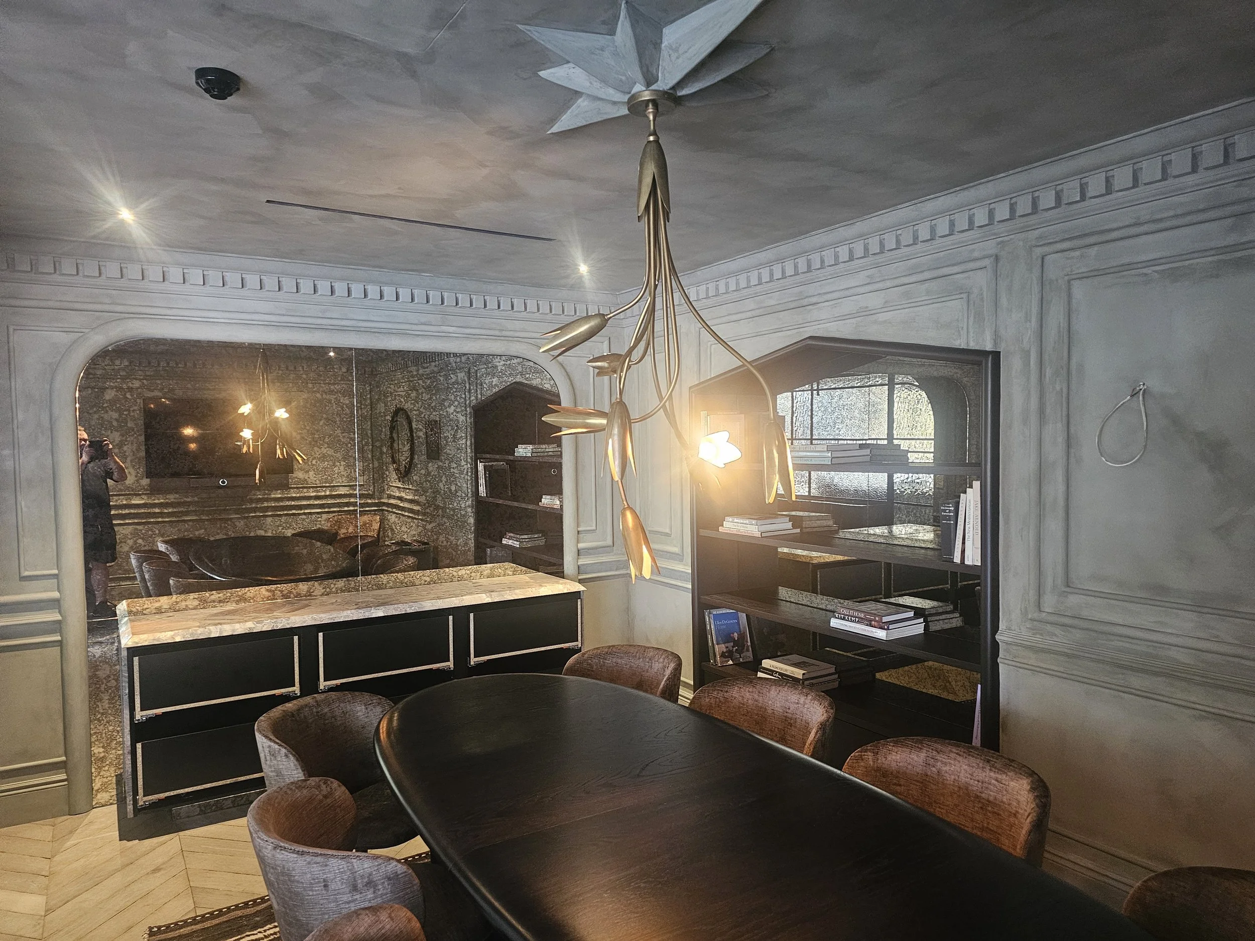 Dining room with a dark wooden table surrounded by upholstered chairs, a decorative chandelier hanging from the ceiling, a black bookshelf with books, and a large mirror on the wall reflecting a television and a person taking the photo.
