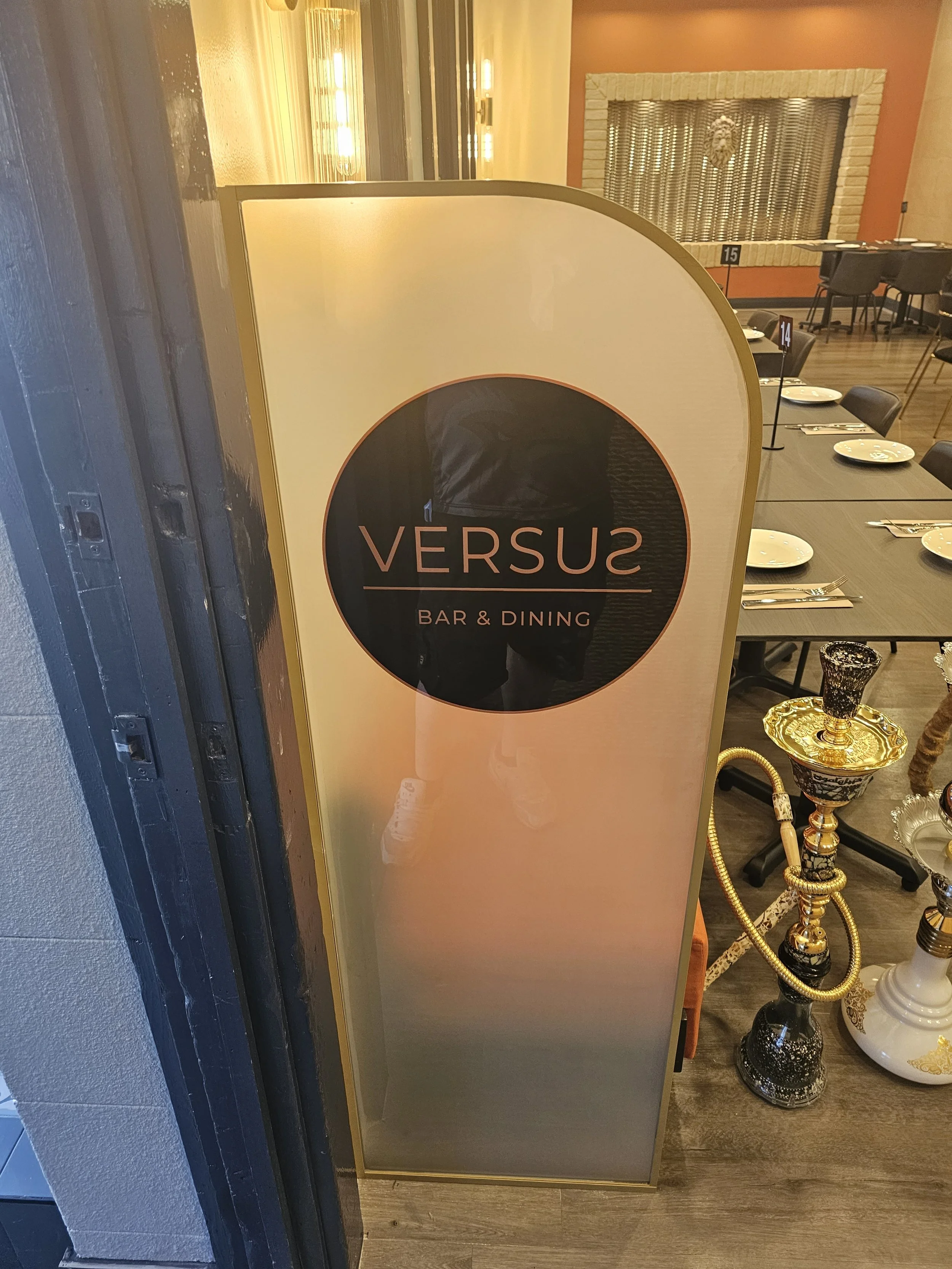 Sign for Versus Bar & Dining, located next to a dining area with tables set with plates and utensils, and hookah pipes displayed nearby.
