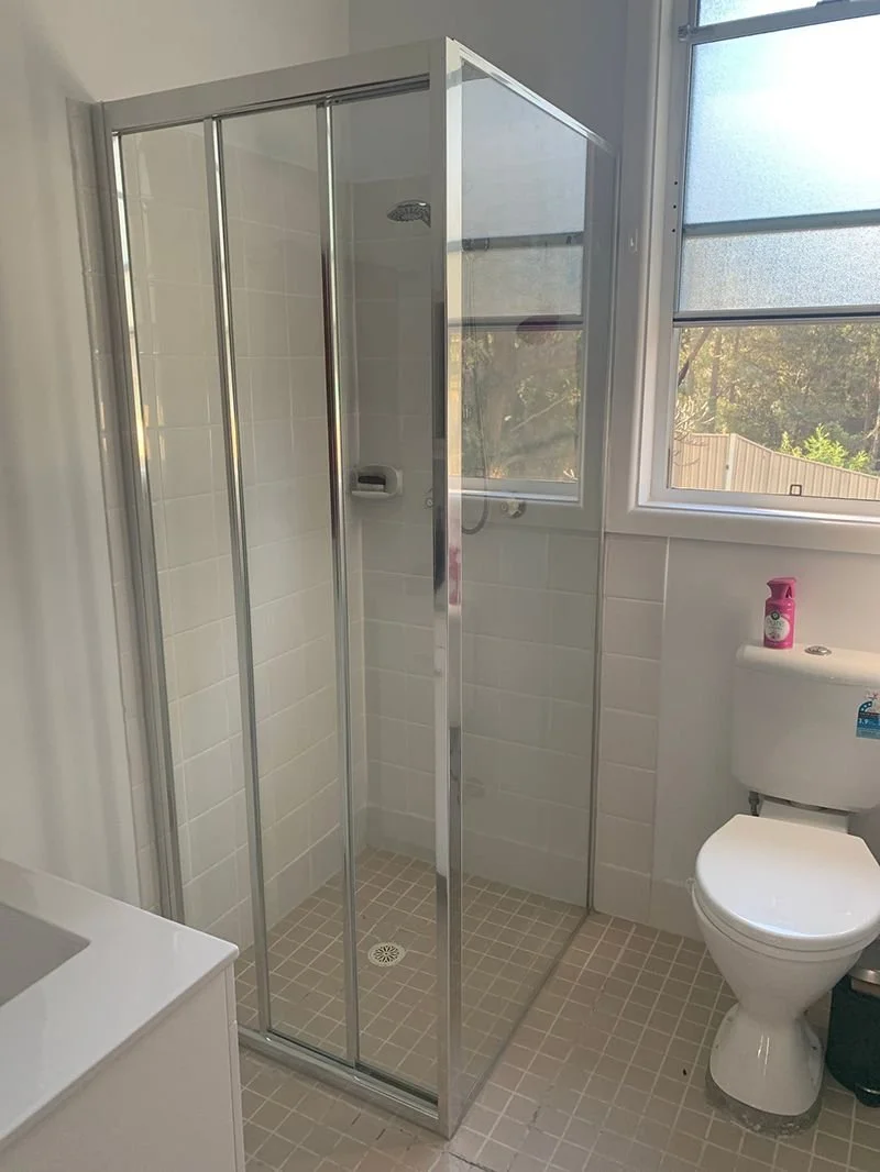Modern bathroom with a walk-in shower enclosed by glass, featuring a rainfall showerhead and handheld shower fixture with gold accents. A wooden vanity with a vessel sink and gold faucet is visible to the right.