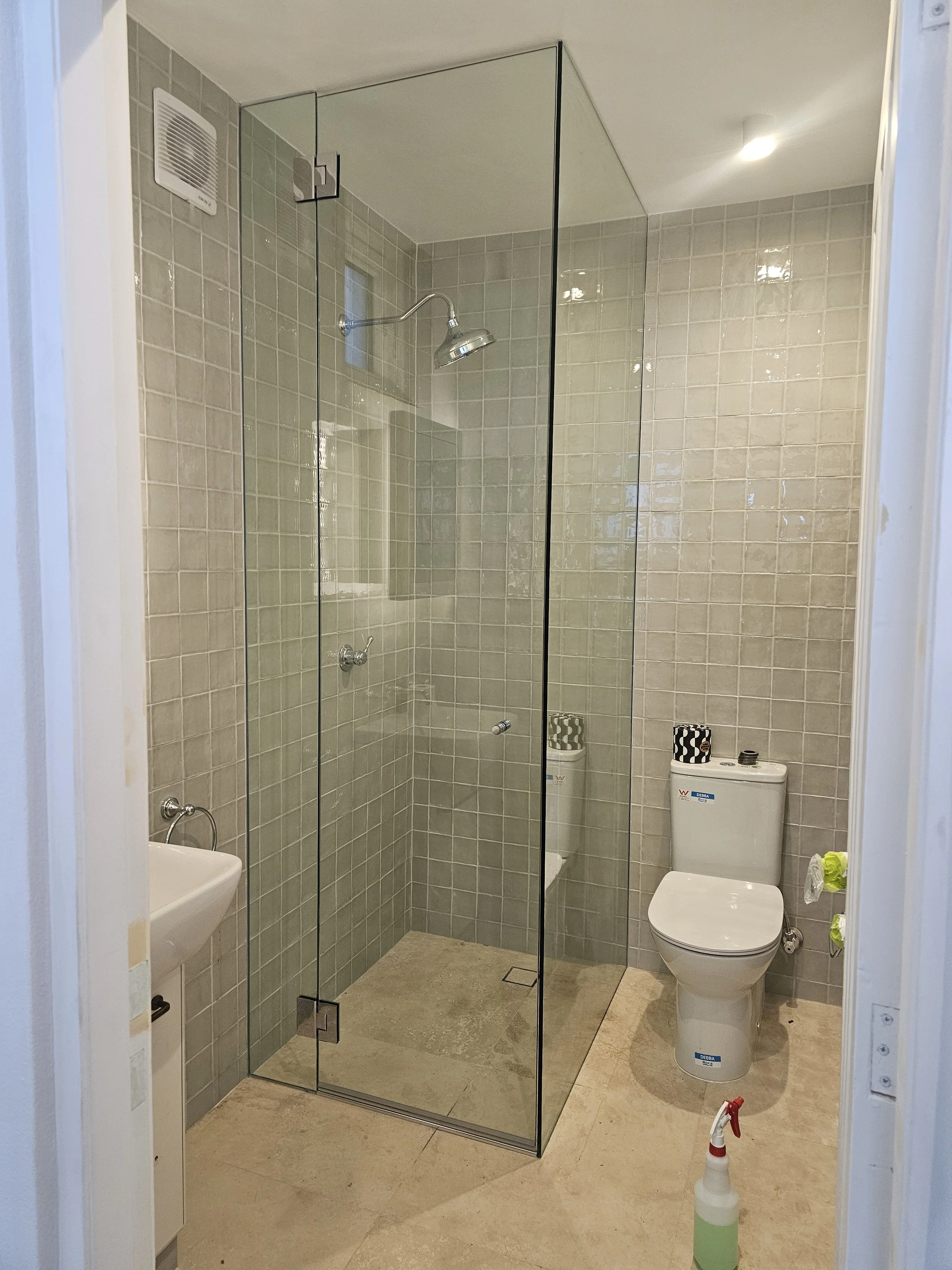 Bathroom with a glass-enclosed shower, a toilet, a small sink, beige tiled walls, and a spray bottle on the floor.