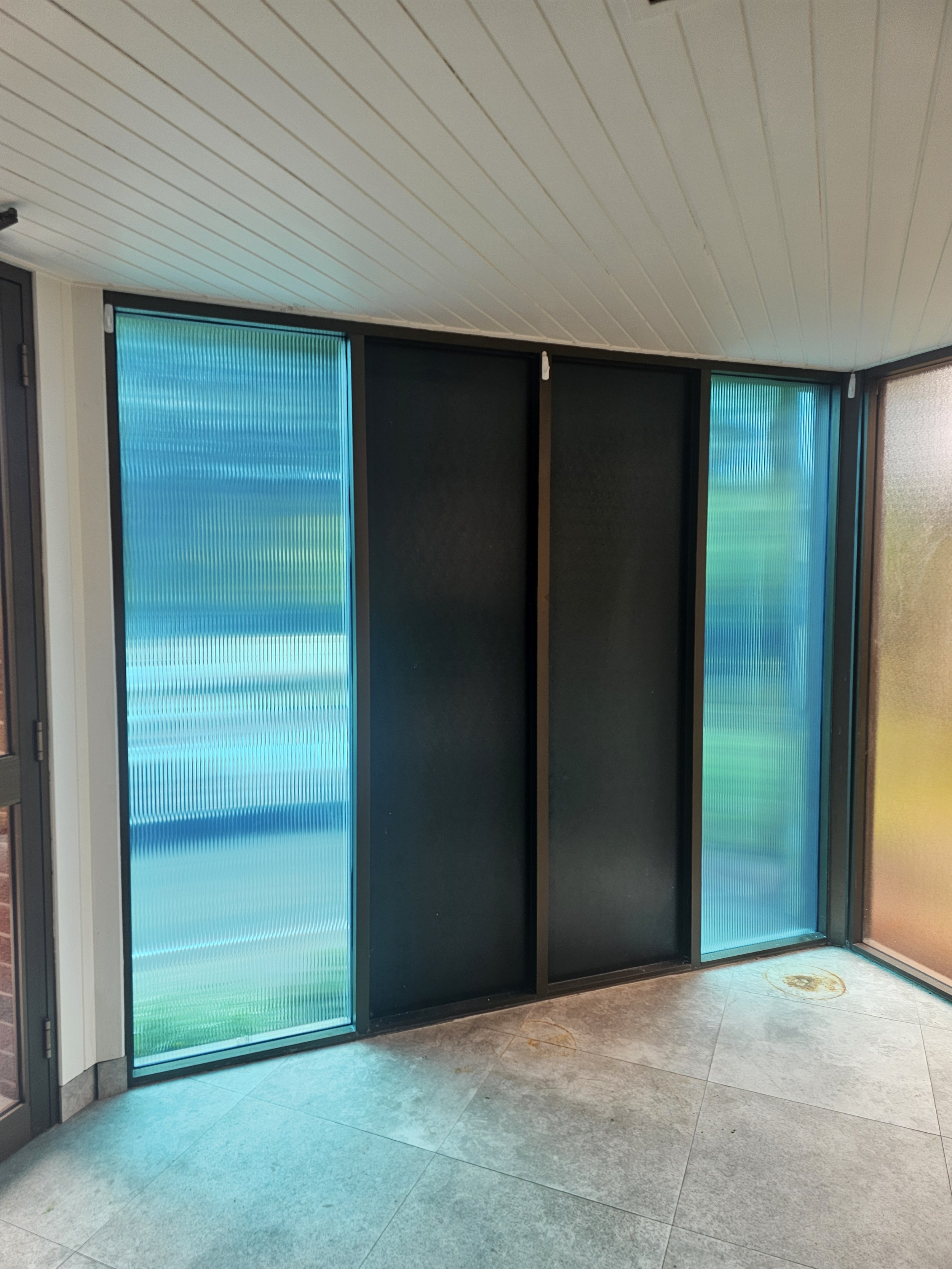 Interior view of a corner with sliding doors, one made of black panels and the others of translucent blue-green textured glass, on a tiled floor with a white wood-paneled ceiling.
