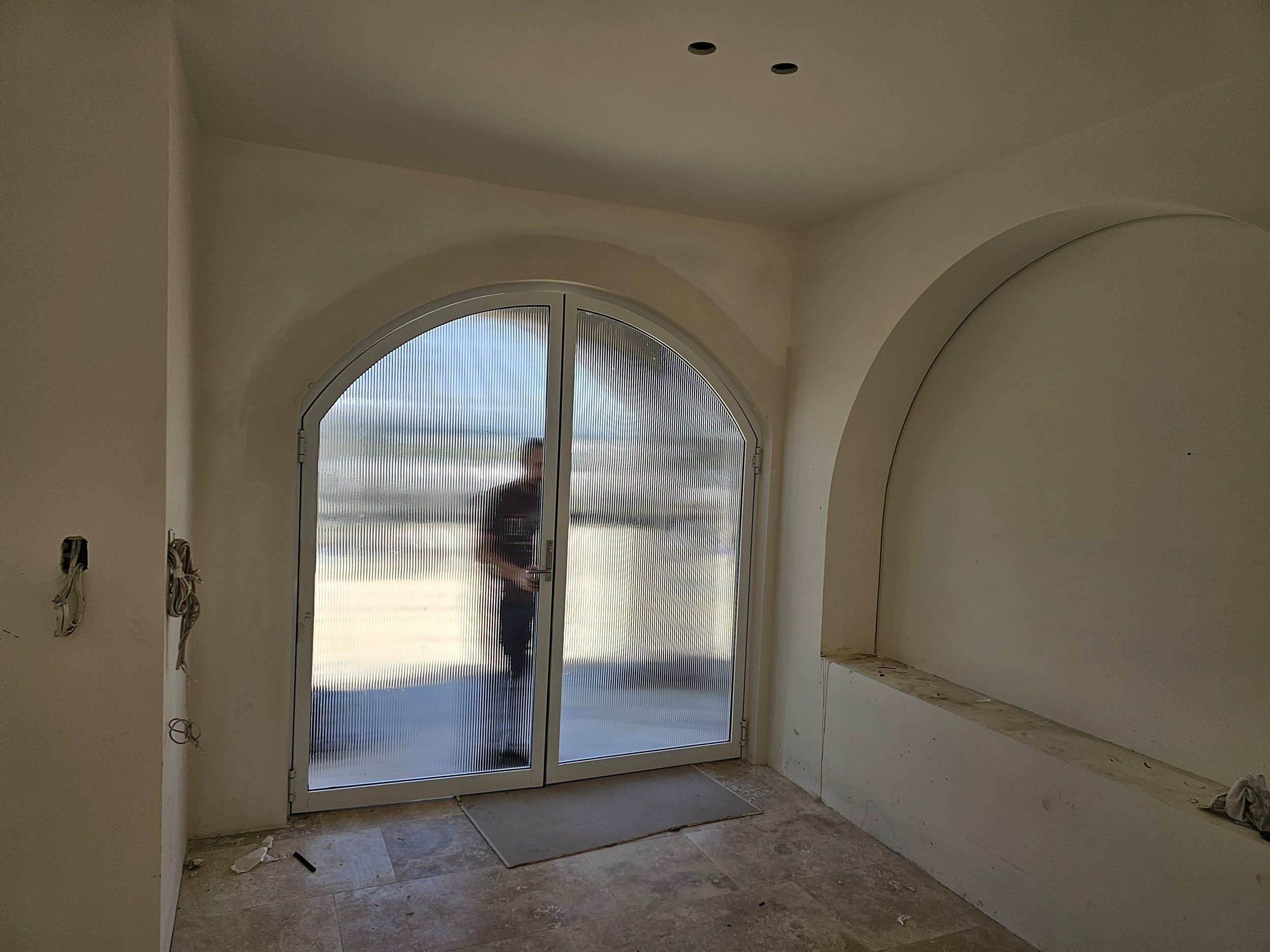 A doorway with frosted glass panels and an arched top, with a person standing outside visible through the glass, in an unfinished room with a tile floor and built-in wall niches.