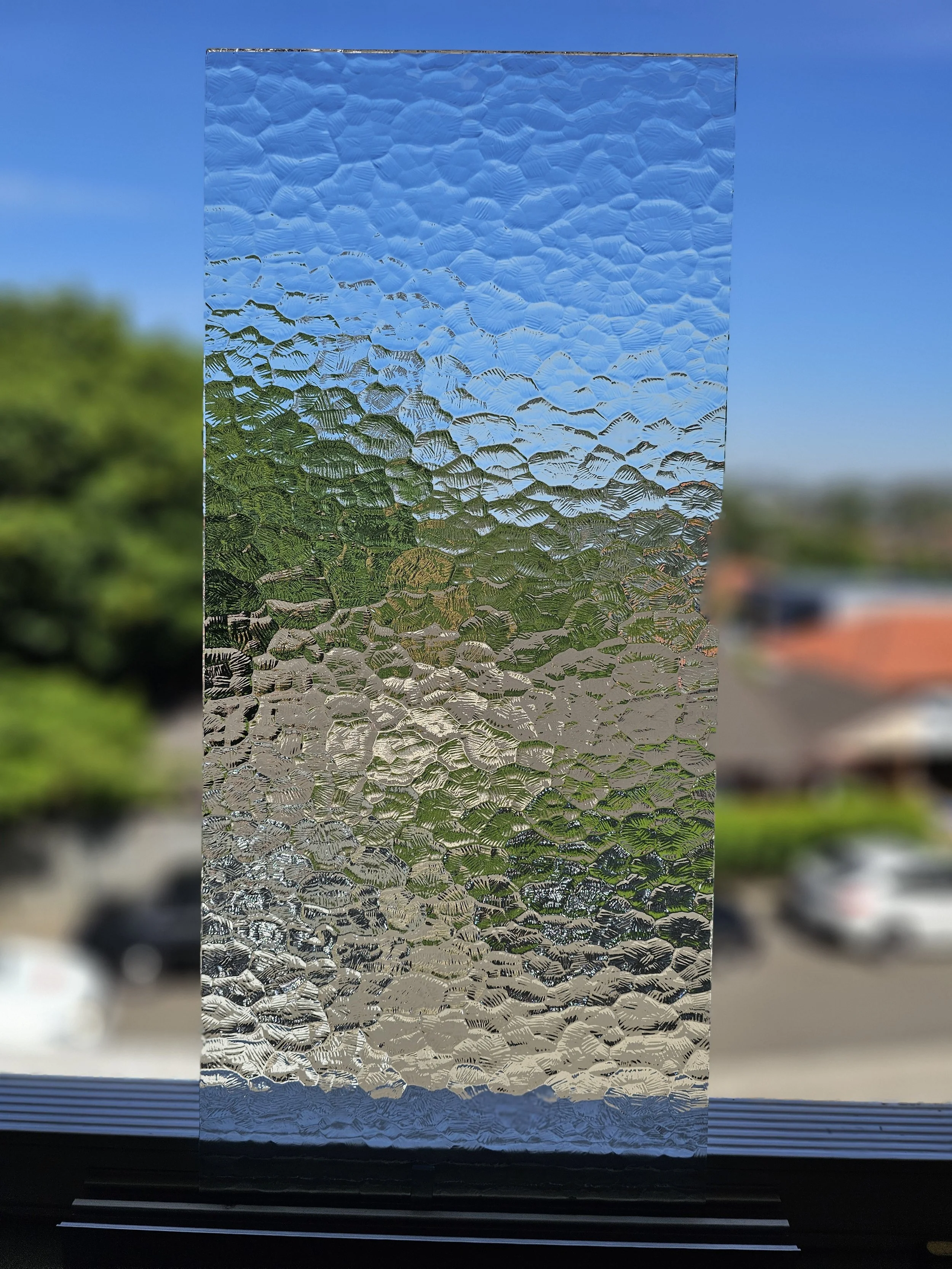 Close-up of textured glass window with a blurred outdoor scene in the background, including trees, parked cars, and a building.