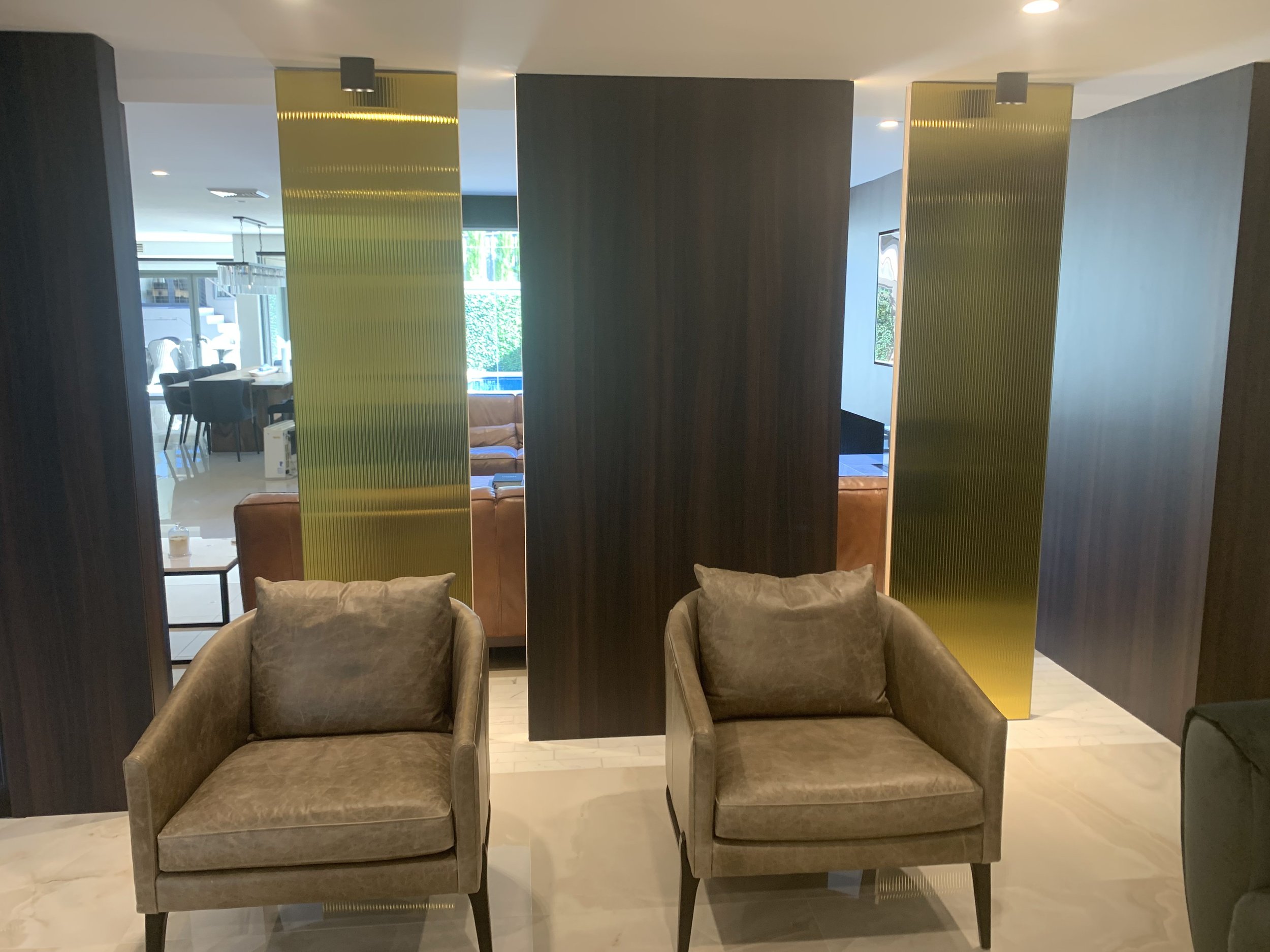 Two beige armchairs with cushions in front of a modern decor wall with dark wood and gold metallic panels, with a view of a dining area and large windows in the background.