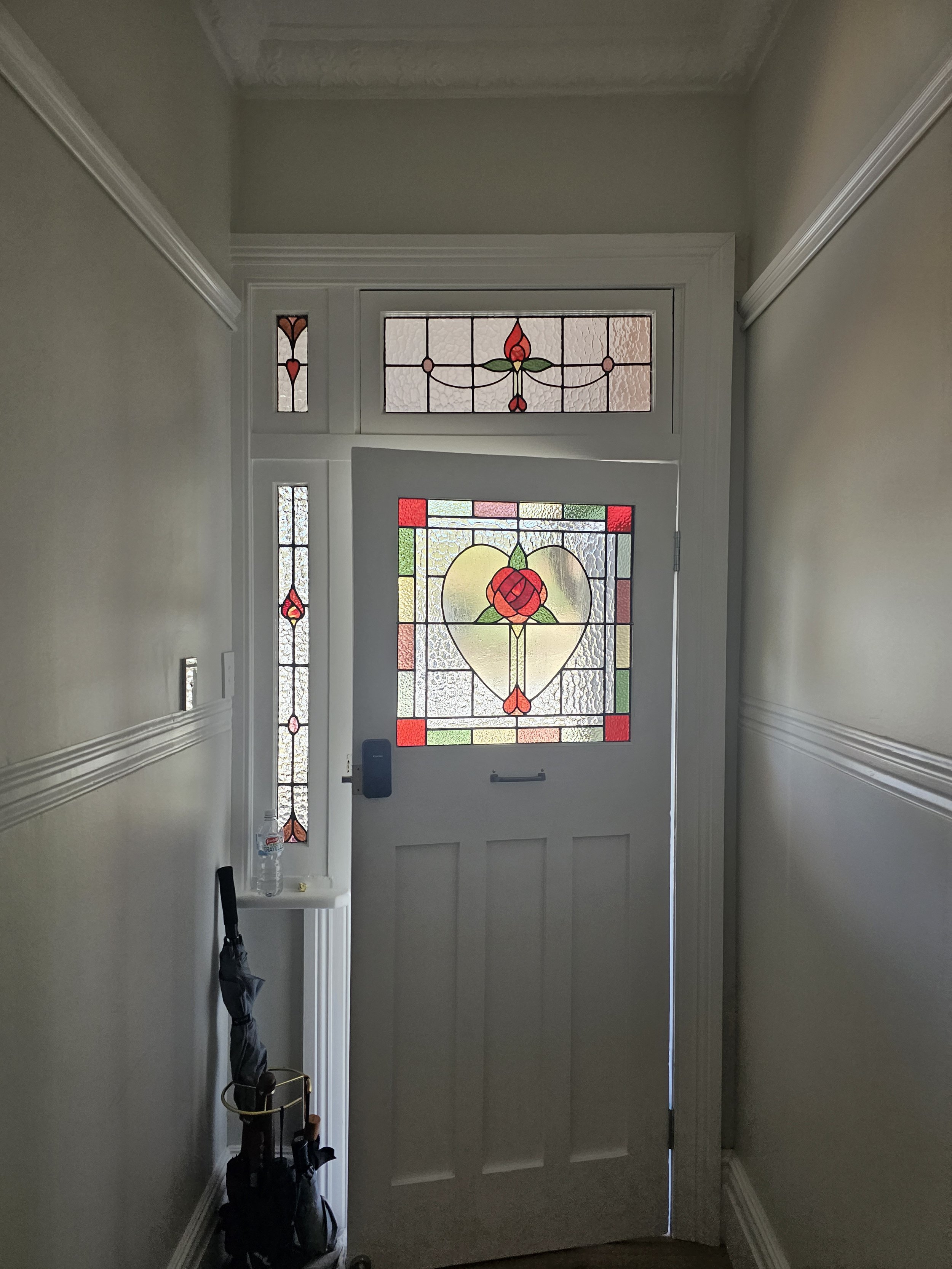 Front entry door with decorative stained glass, featuring a heart and floral design, flanked by narrow stained glass panels, in a hallway with light-colored walls and crown molding.