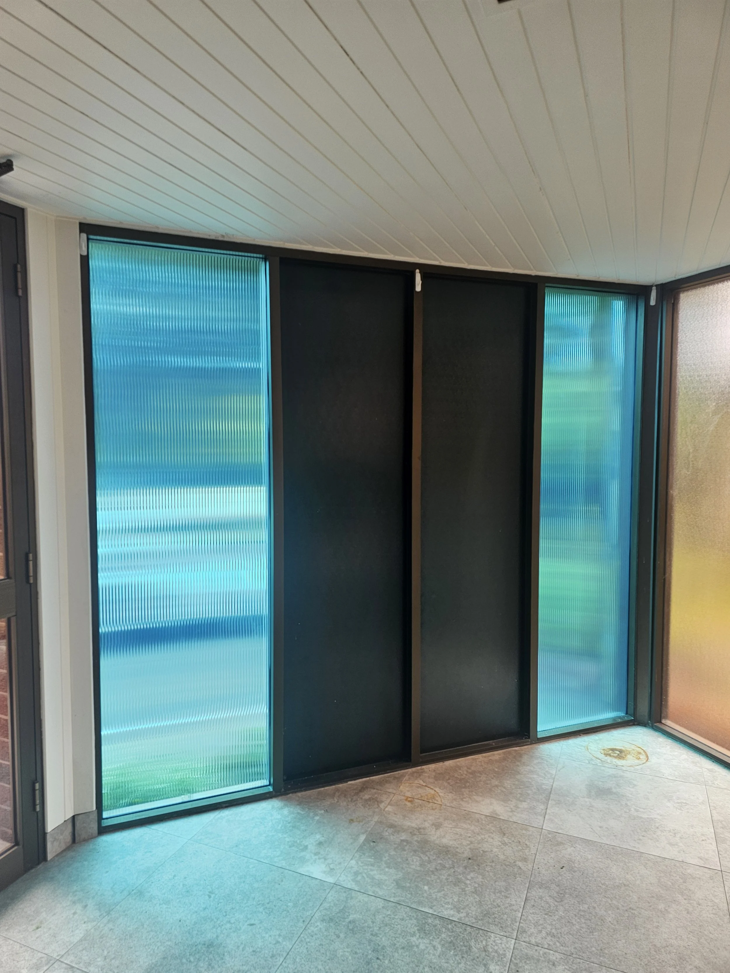 A corner of an indoor space with sliding black doors, two with translucent blue-green textured panels, set against a white wooden-paneled ceiling and tiled floor.