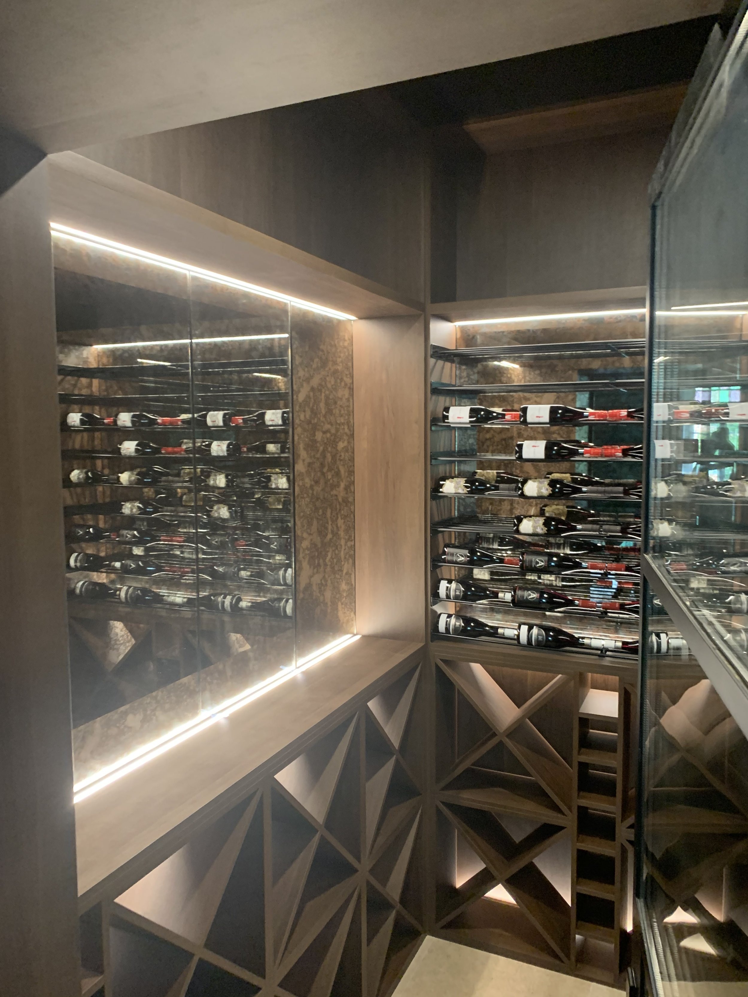 A modern wine storage room with wooden shelves and glass doors, holding bottles of wine, illuminated by built-in lighting.