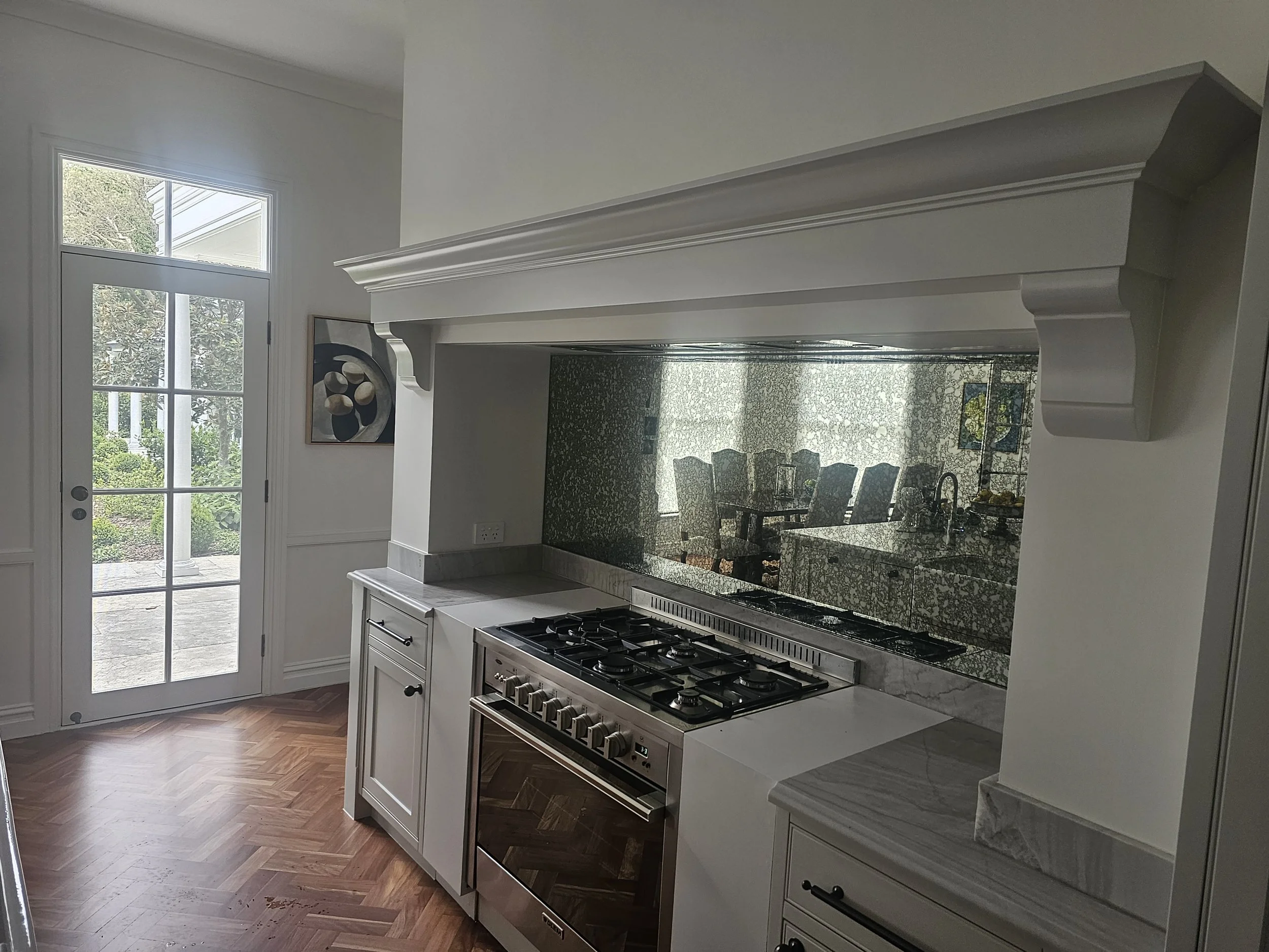 Kitchen with white cabinetry, marble countertops, and a stainless steel oven. A reflective backsplash extends from the countertops to the ceiling, reflecting a dining area with chairs and a window. A glass door leads outside to a patio, and there's a piece of artwork on the wall.