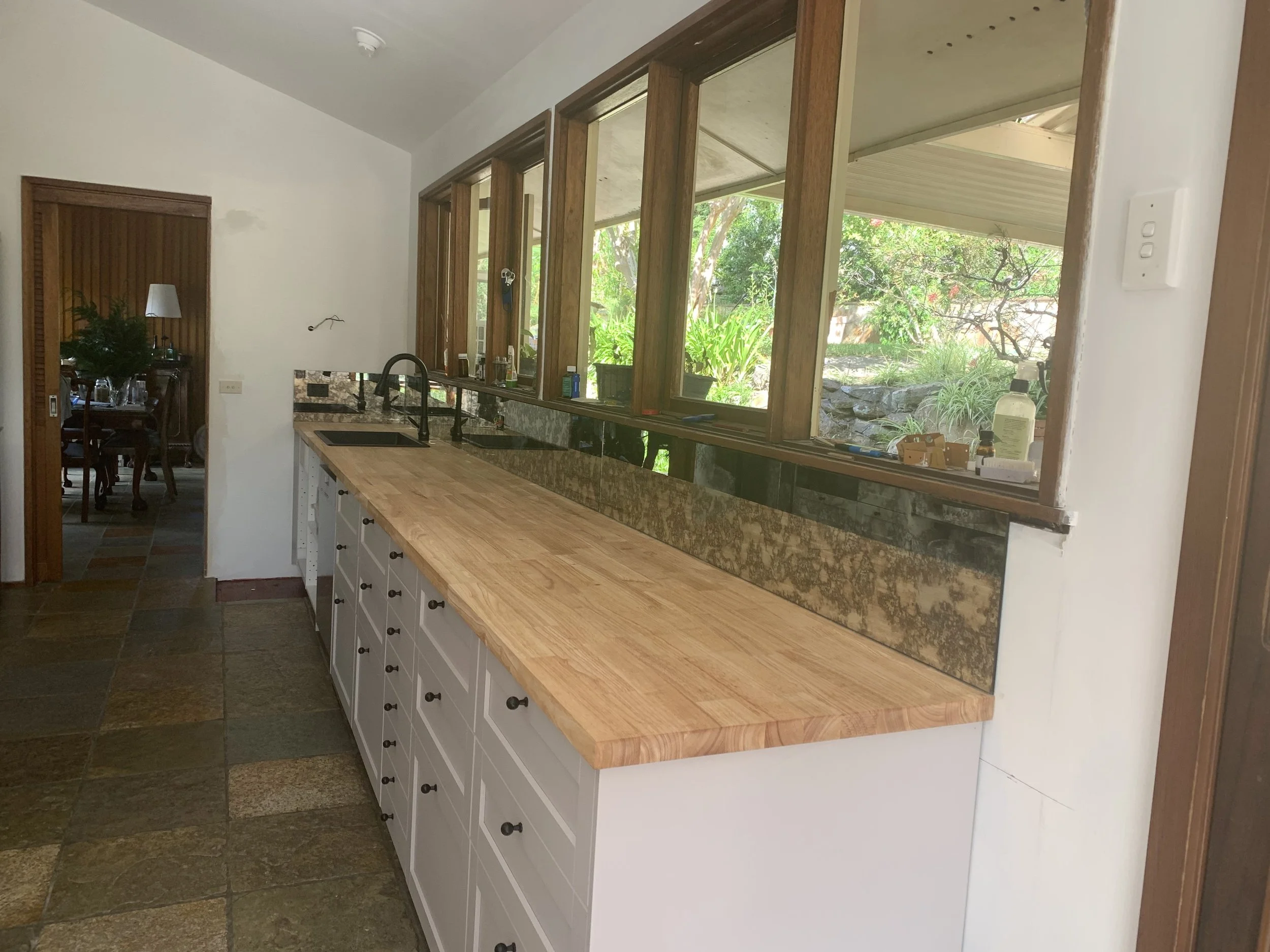 Kitchen with a wooden countertop, white cabinets, black faucet, overlooking a garden through large window openings.