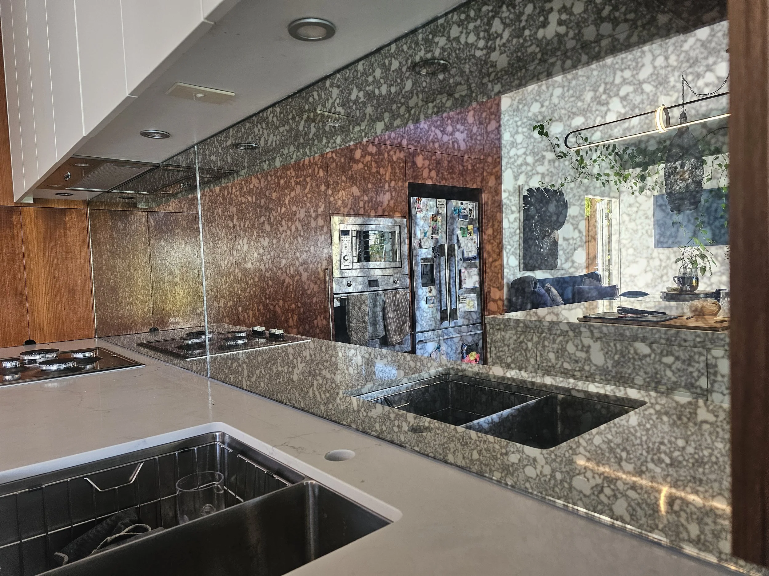 Kitchen with a reflective backsplash showing a living room with a couch, window, hanging light fixture, plants, and kitchen appliances.