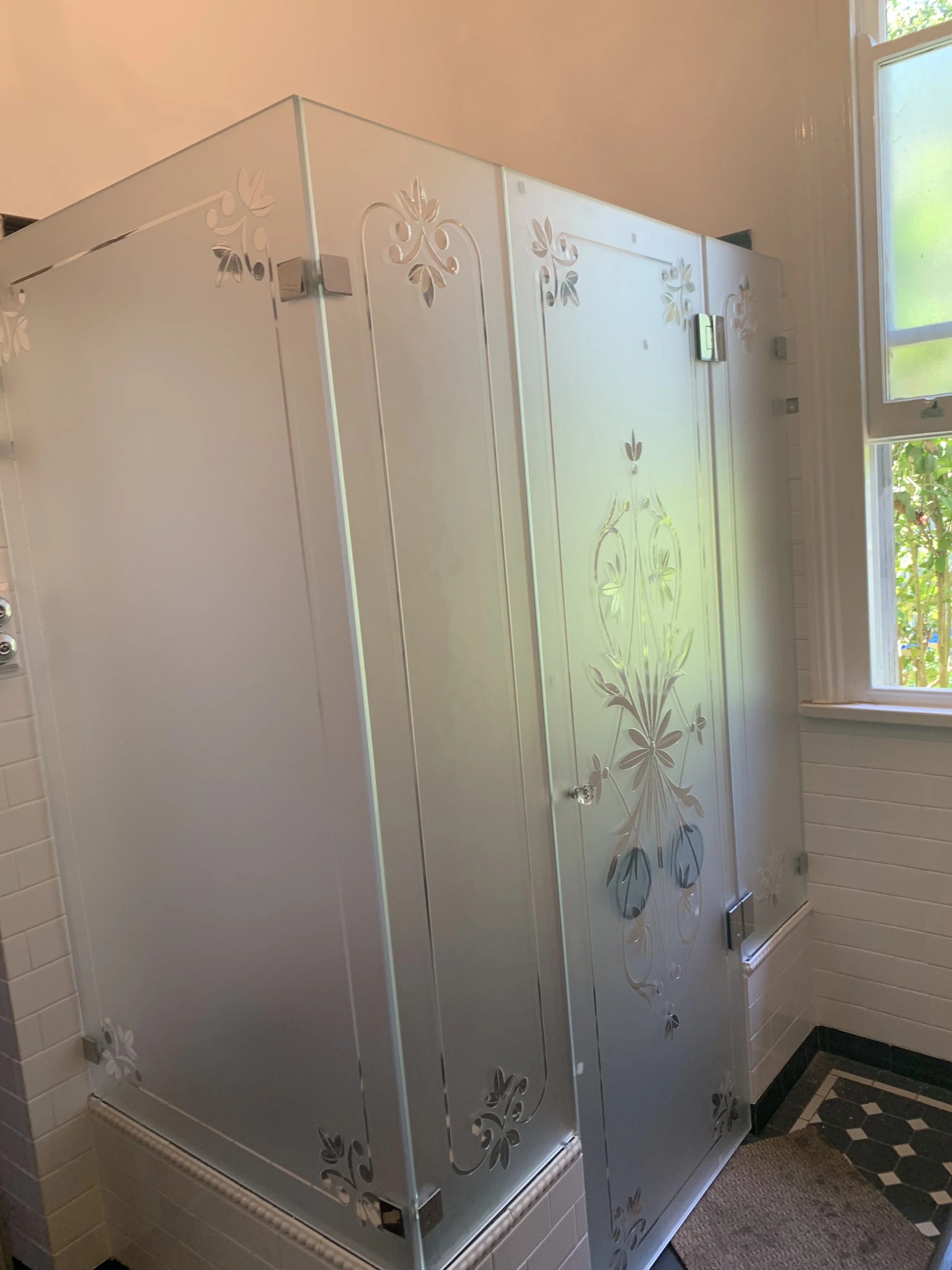 A vintage shower with frosted glass panels decorated with floral patterns, located in a tiled bathroom near a window.