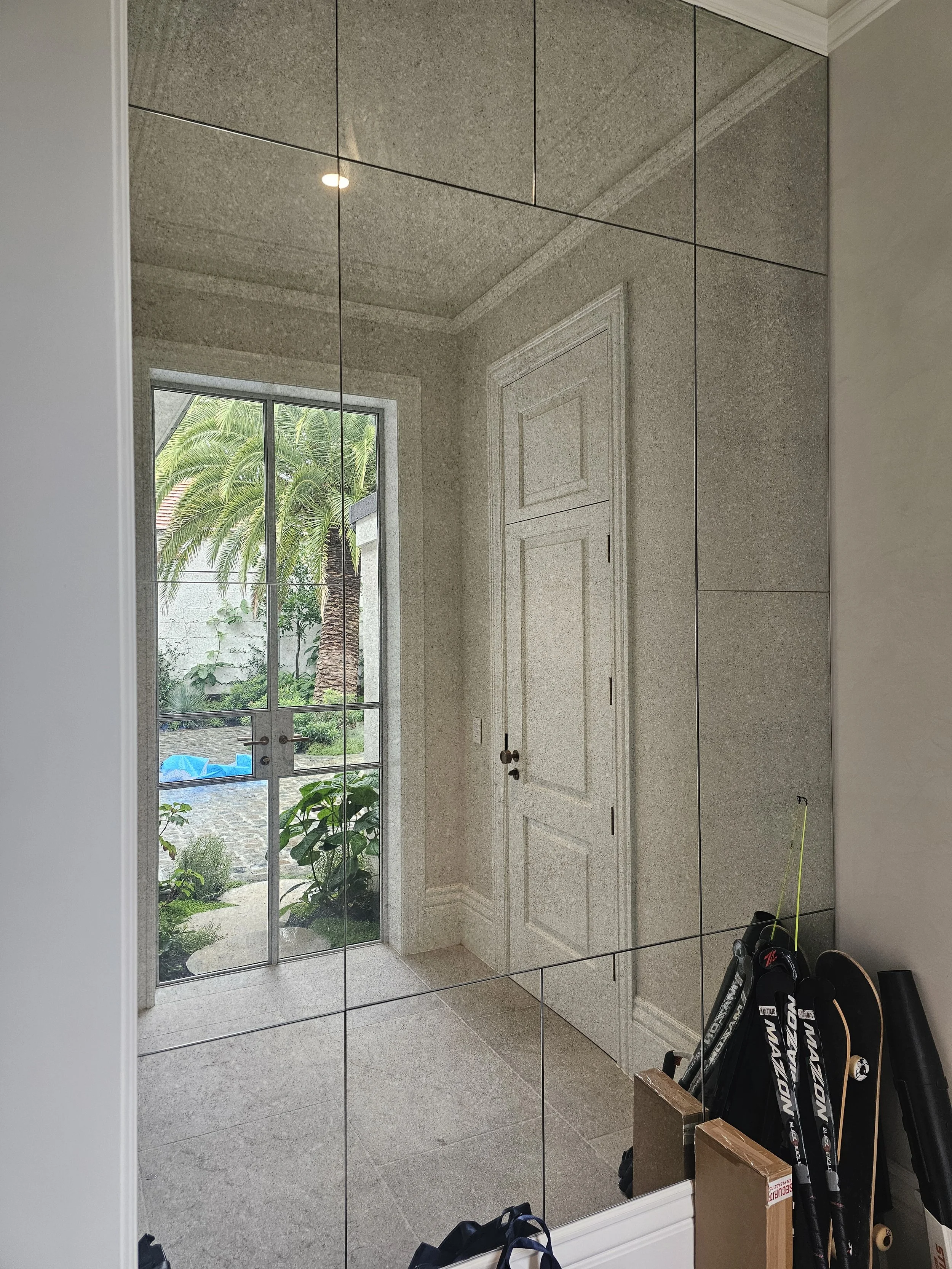 A large mirror in a hallway reflects a white door, a window showing palm trees outside, and some sports equipment including skis and a skateboard.