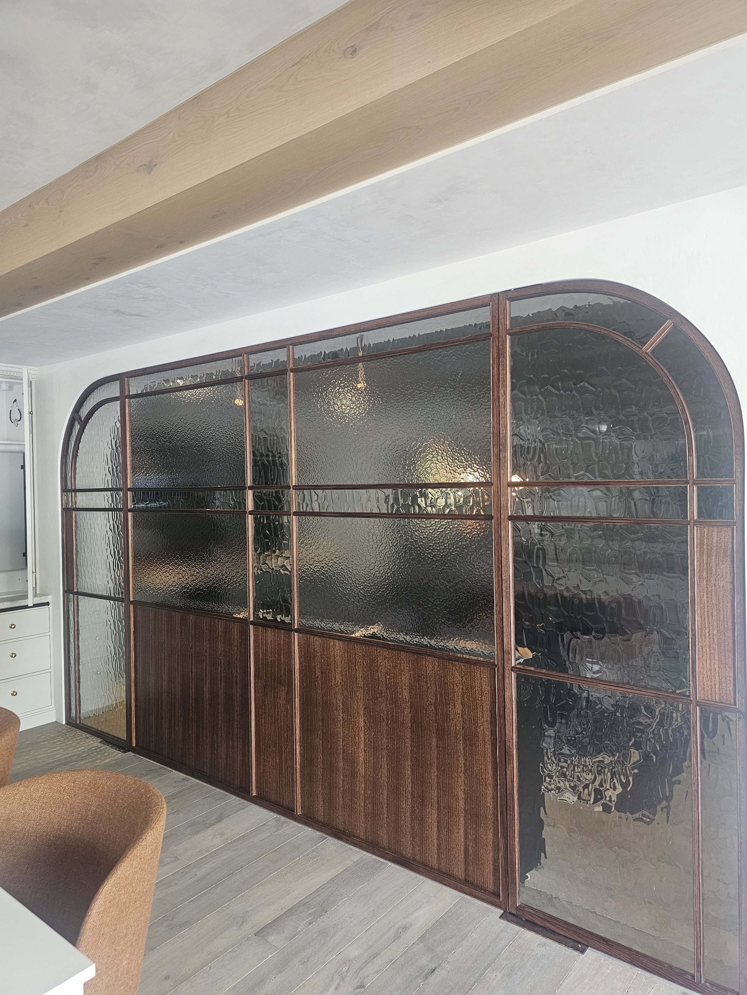 A room divider with wooden frame and textured glass panels, featuring an arched top on the right side and rectangular sections, situated next to a white wall and partial view of a chair.