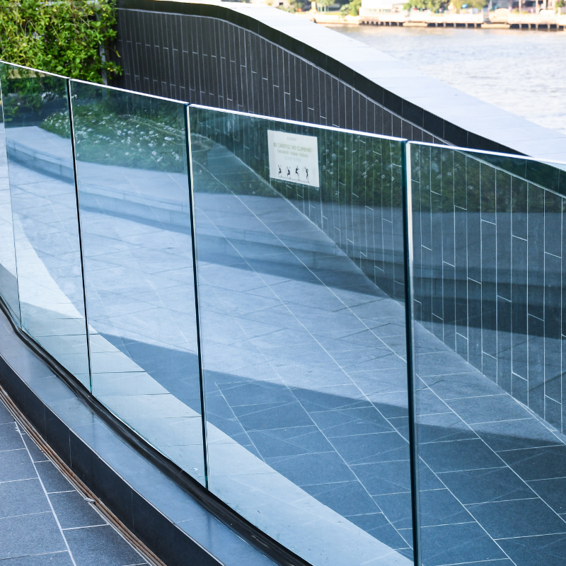 A curved glass railing along a waterway, with a black tiled wall behind it and a sign on the wall. Reflection of the surroundings can be seen in the glass.