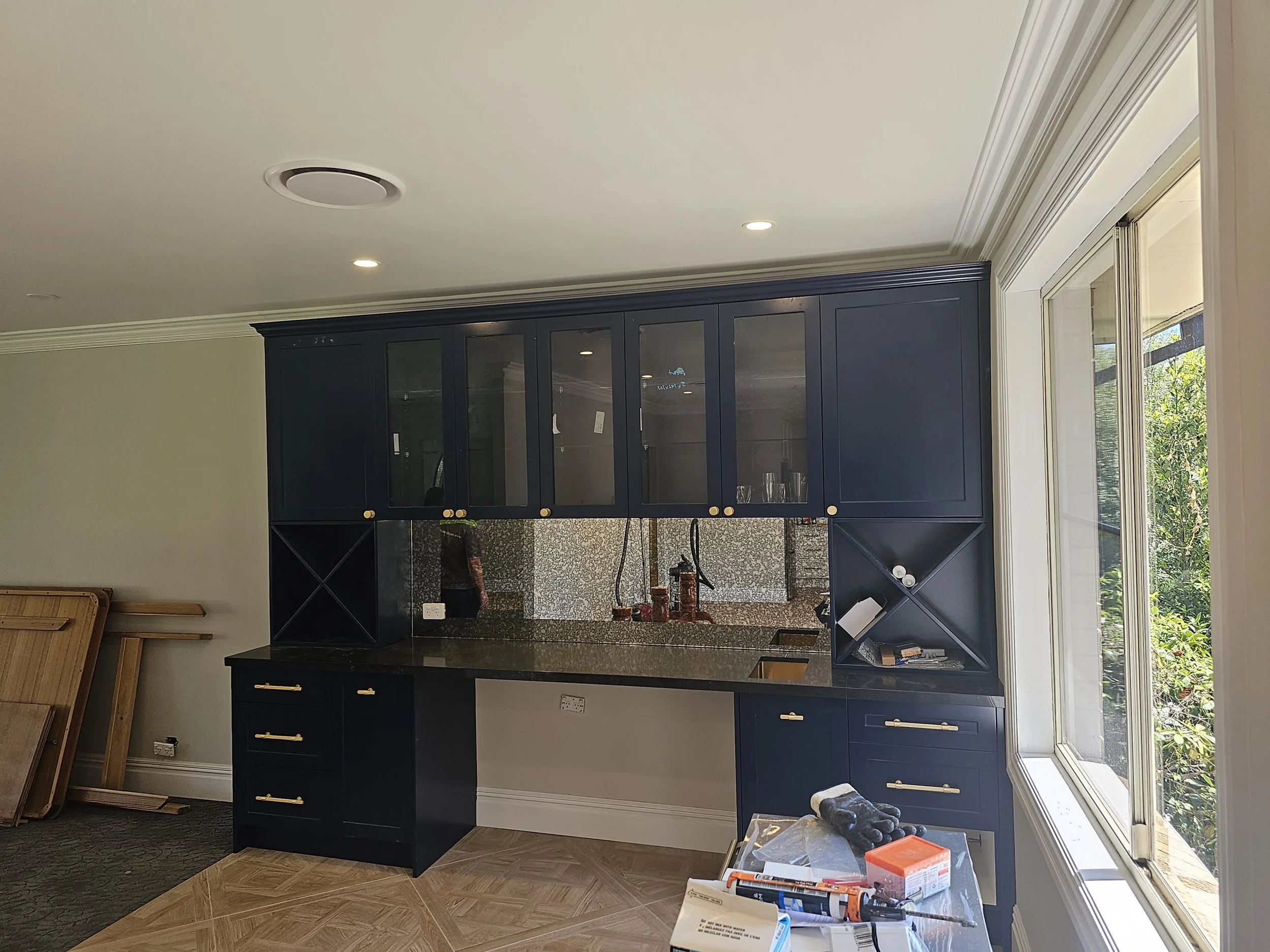 Kitchen cabinet being installed near a large window, with construction tools and materials on a table in the foreground.